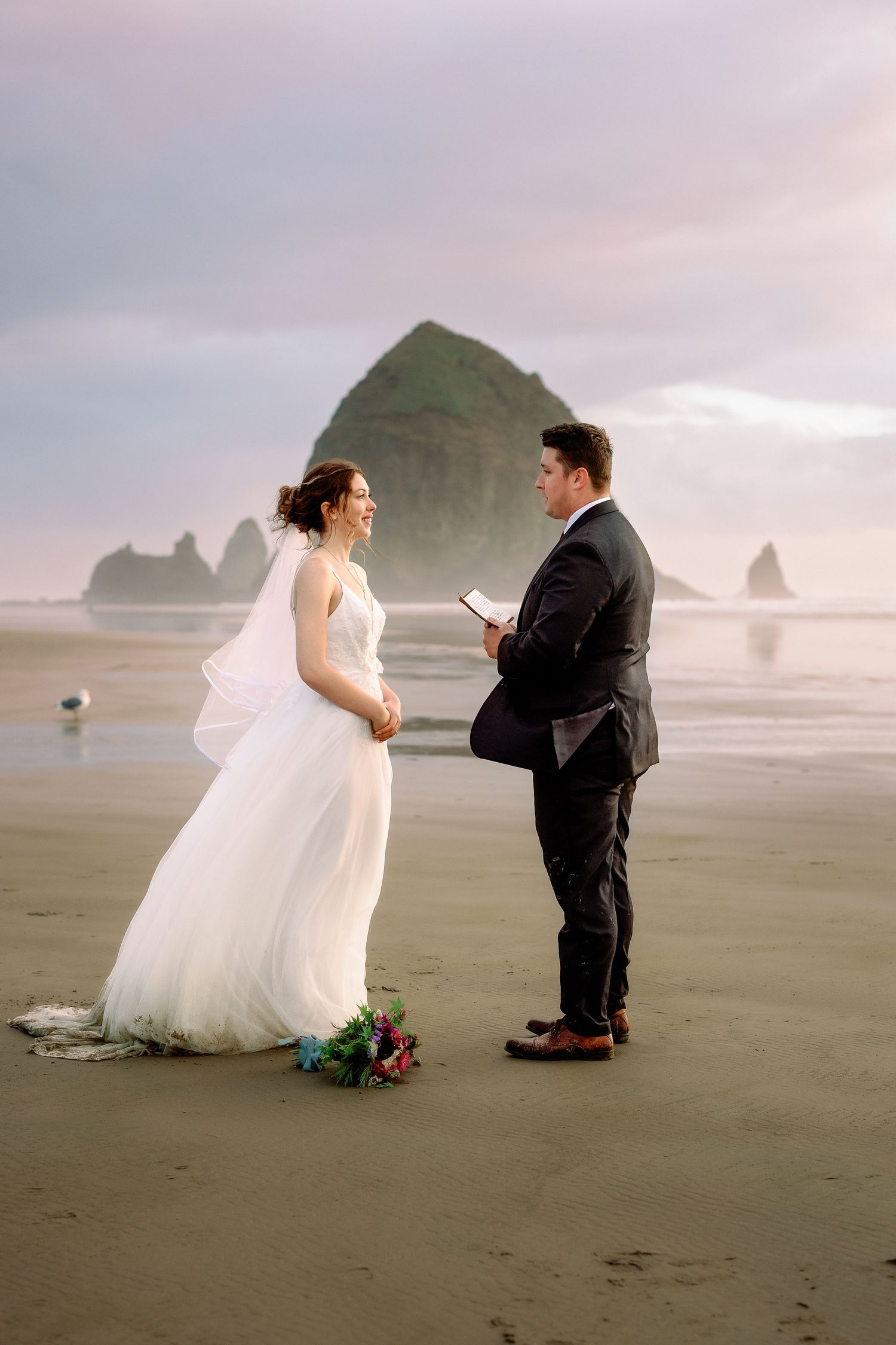 A couple shares intimate wedding moments on a misty beach with dramatic rock formations in the background.