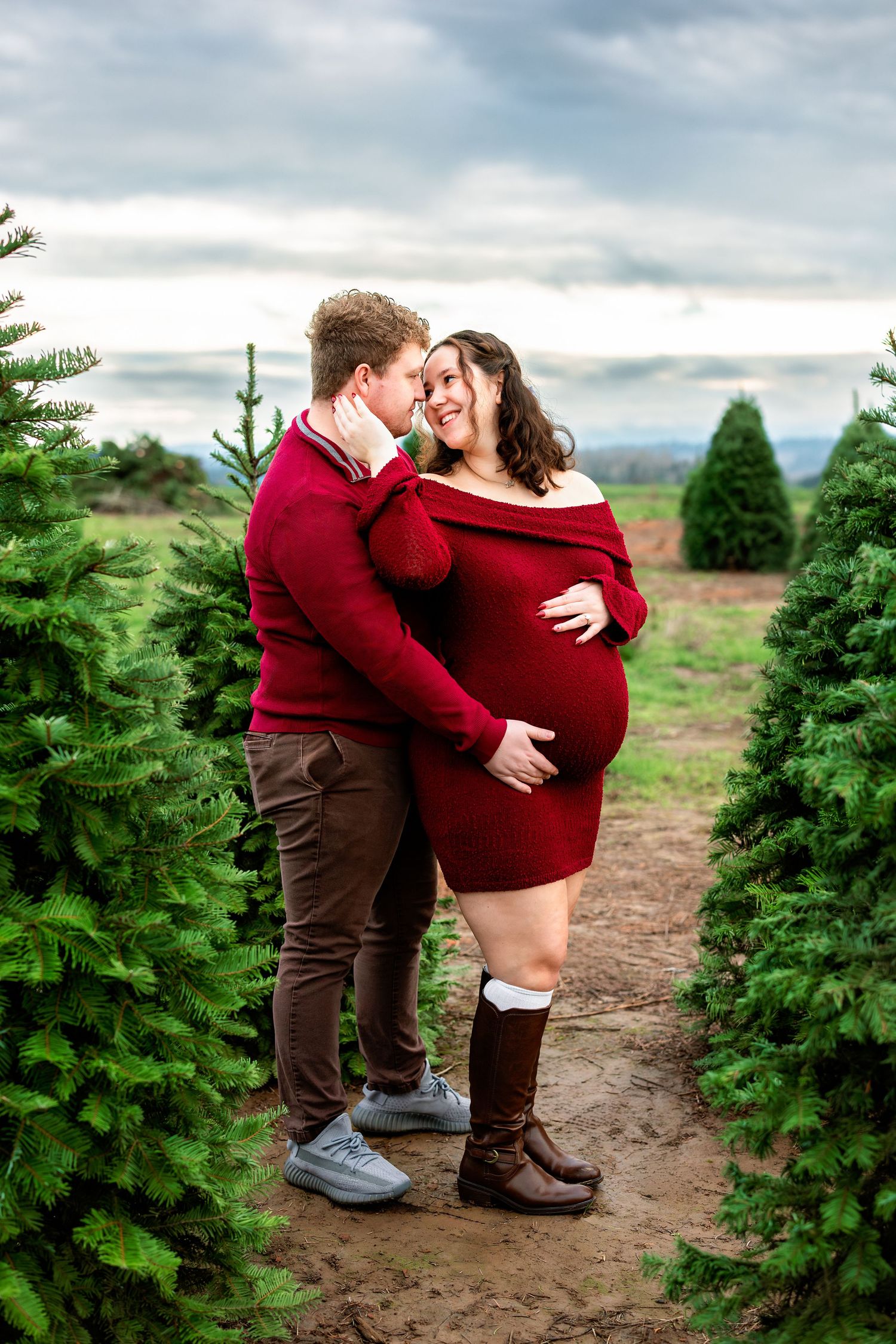 A couple embraces romantically between rows of evergreen trees at a Christmas tree farm on an overcast day.