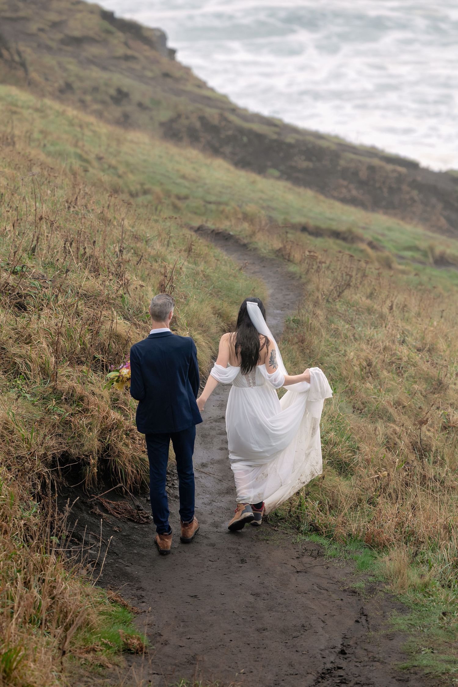 A couple walking down a scenic coastal trail with long grass and ocean views in the background.