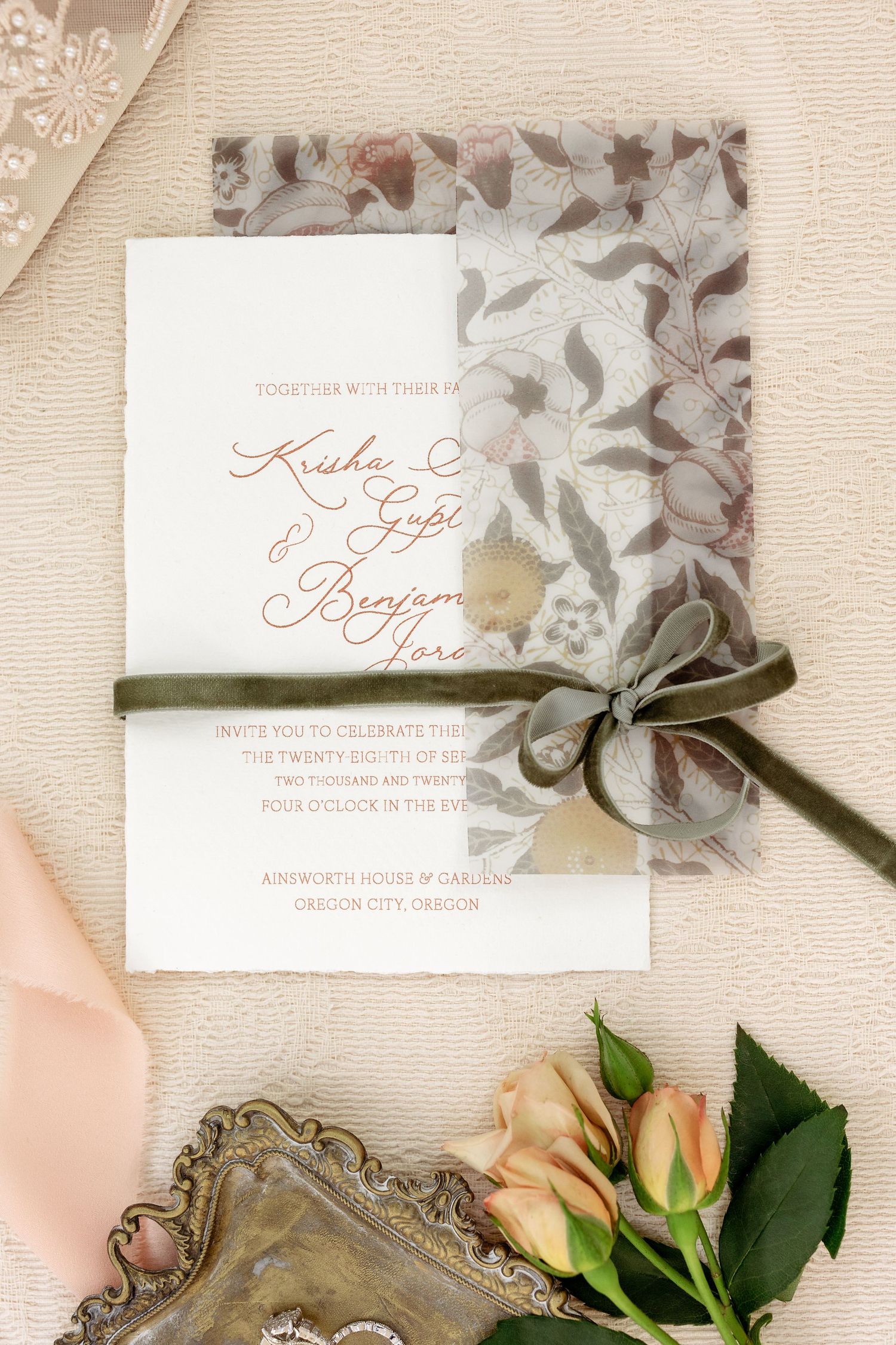 Elegant wedding invitation with floral pattern and sage green ribbon displayed with roses on textured paper.