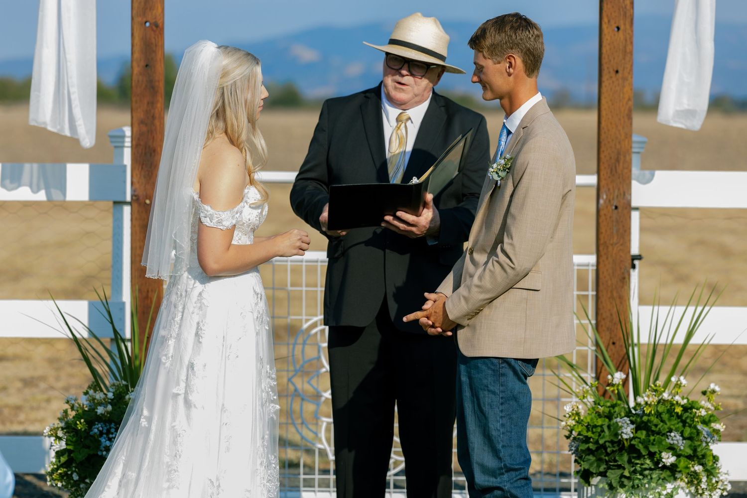 Outdoor wedding ceremony under wooden posts with white fabric draping in a rustic setting.