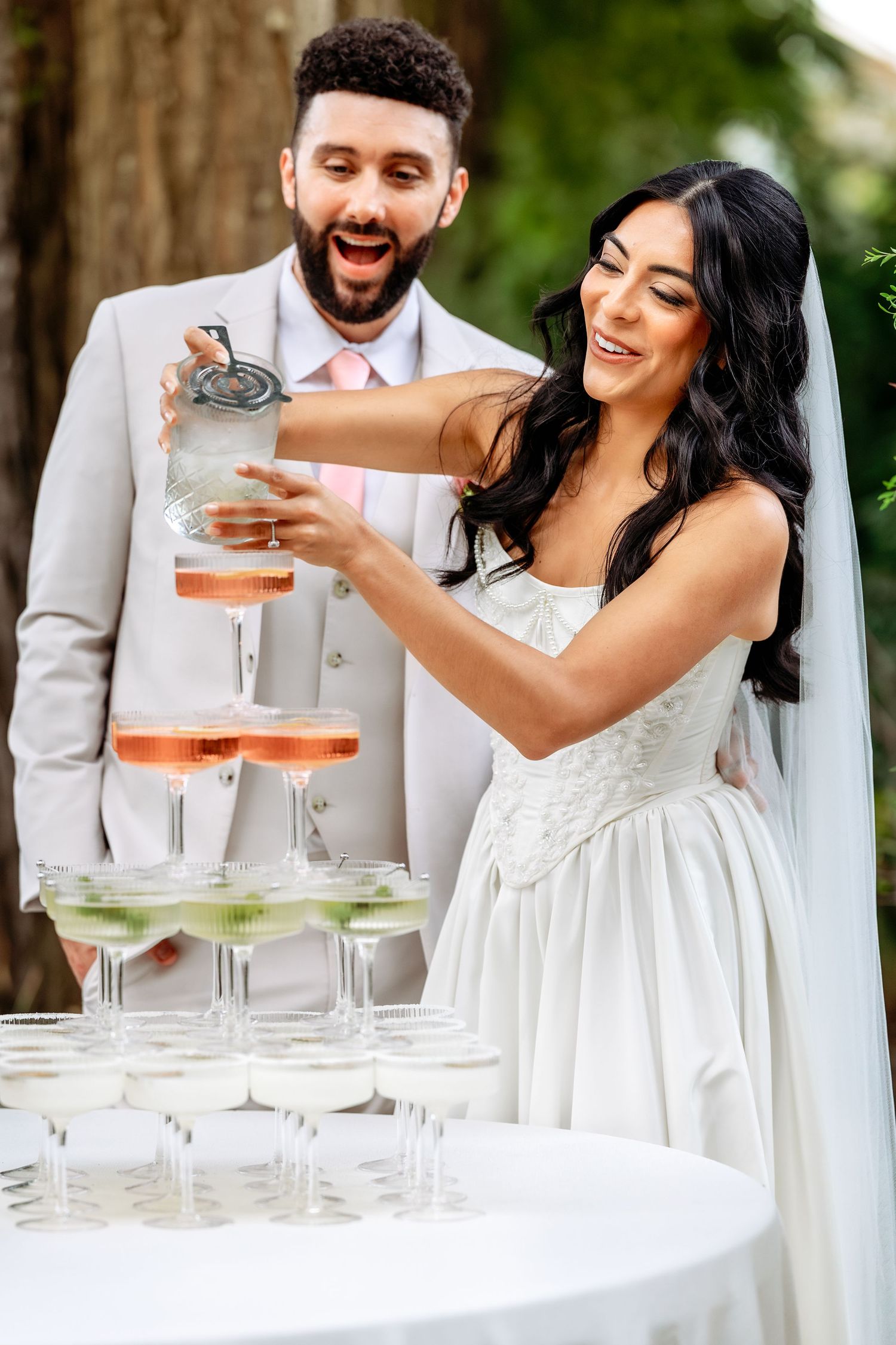 Happy couple pours drinks into a tower of champagne glasses at their outdoor wedding reception.