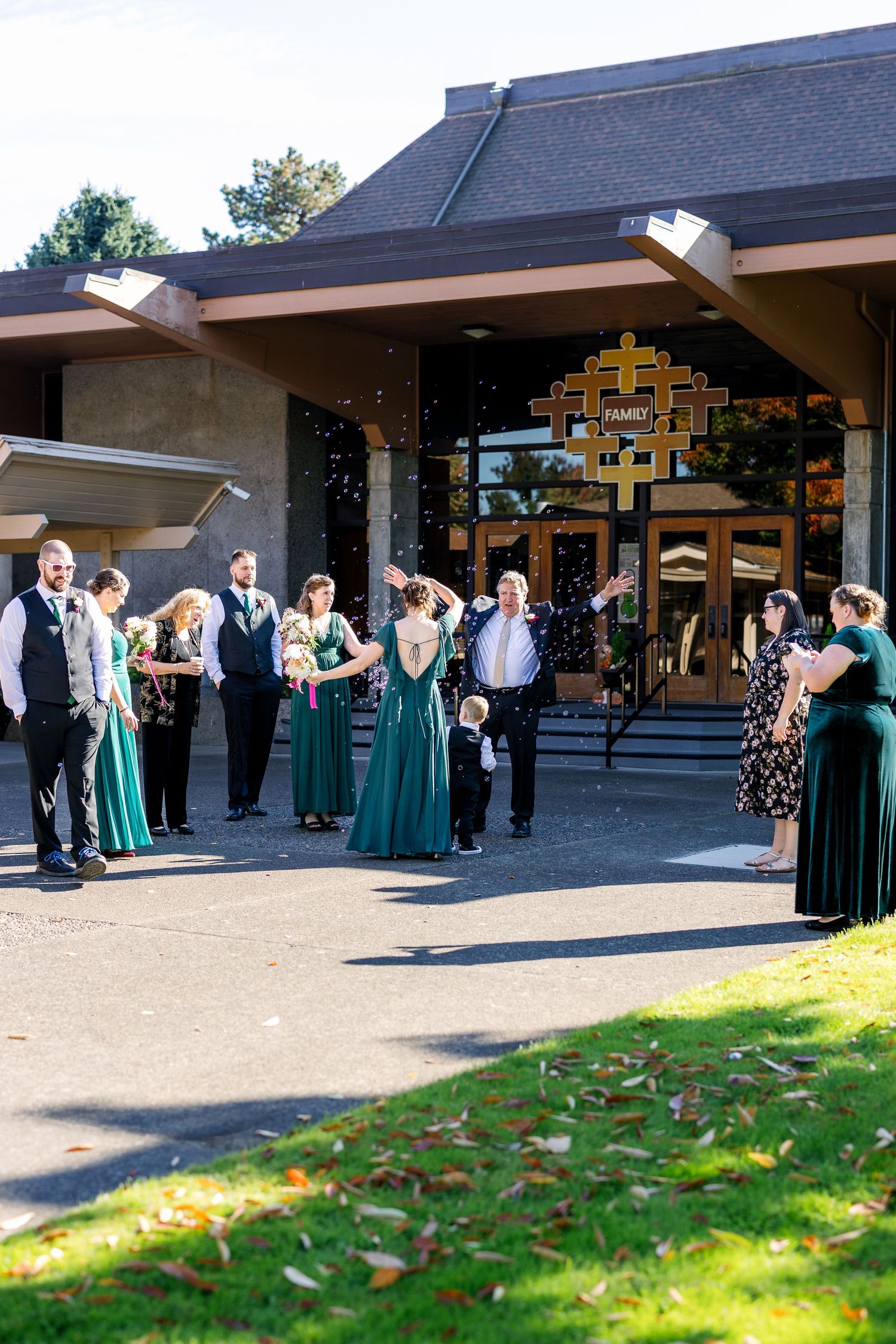 Wedding party members in emerald green dresses and dark suits gather outside a modern church building.