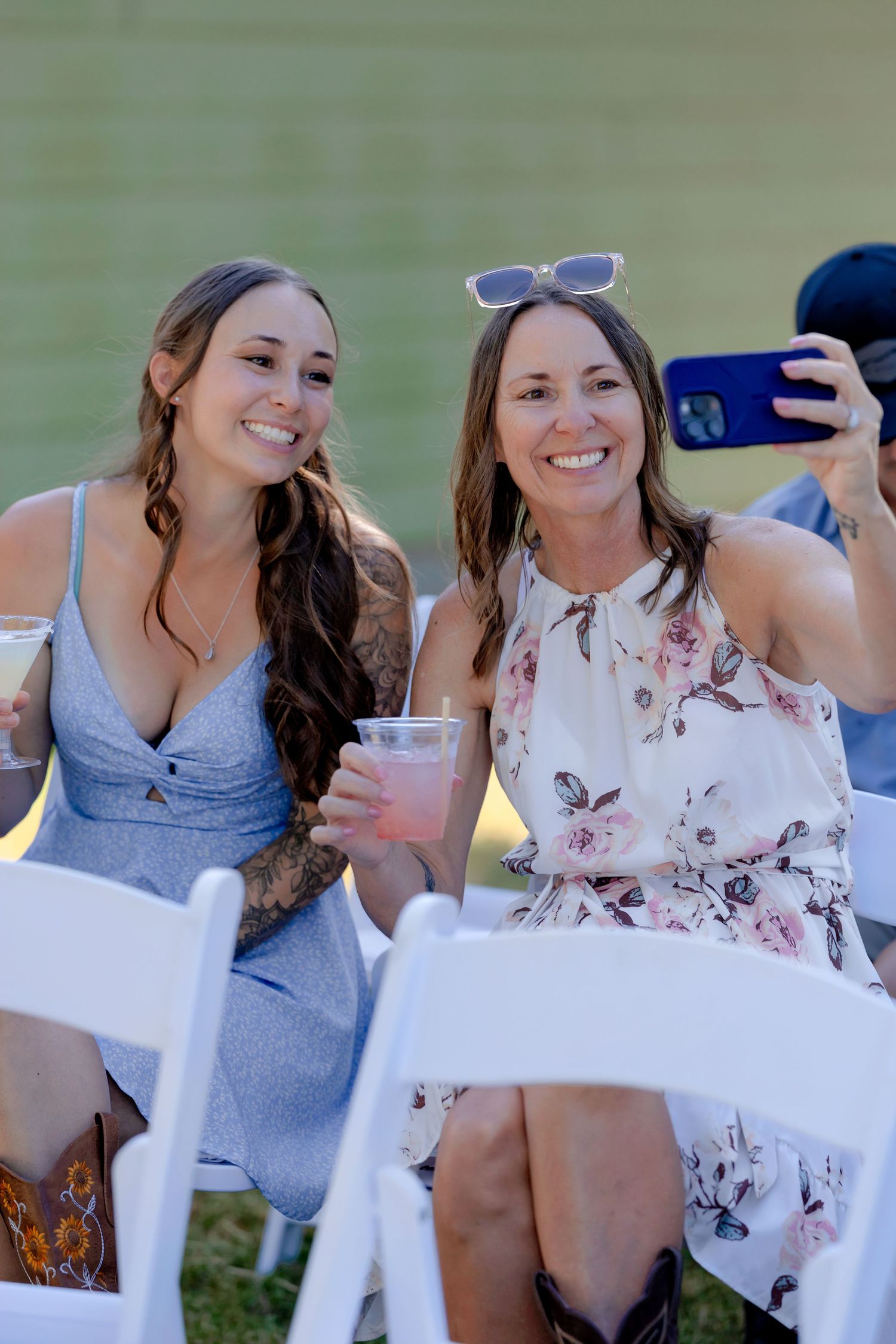 Two smiling women in summer dresses take a selfie while holding drinks at an outdoor wedding reception.