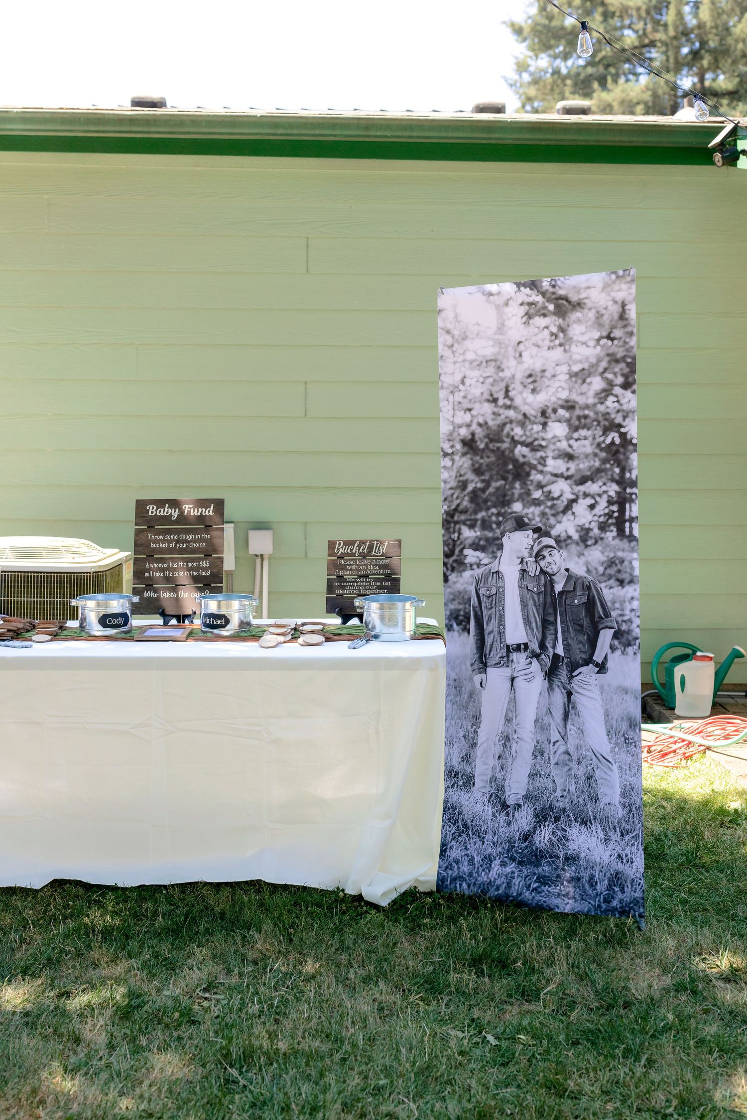 Outdoor wedding welcome table with white tablecloth, decorative signs, and large black-and-white photo banner beside a green building.