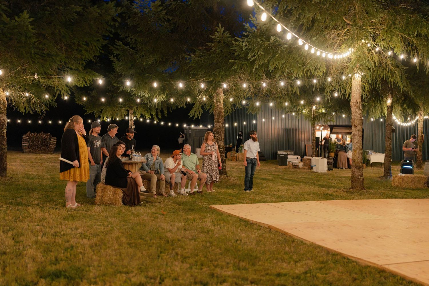 Wedding guests gather under warm string lights on a dance floor at an outdoor evening reception.