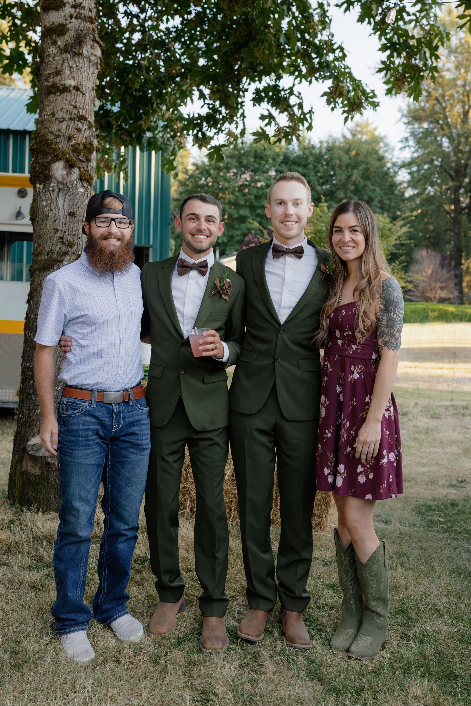 Two grooms in green suits pose with friends outdoors at a rustic wedding venue surrounded by trees.