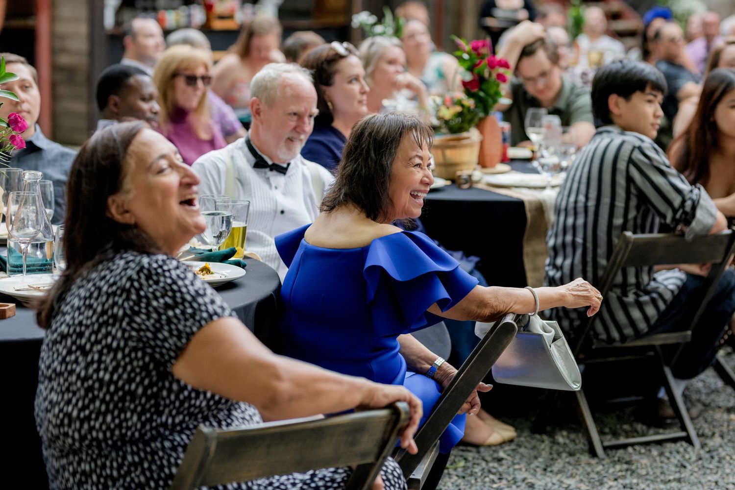 Wedding reception guests laughing and enjoying speeches at outdoor dinner tables.