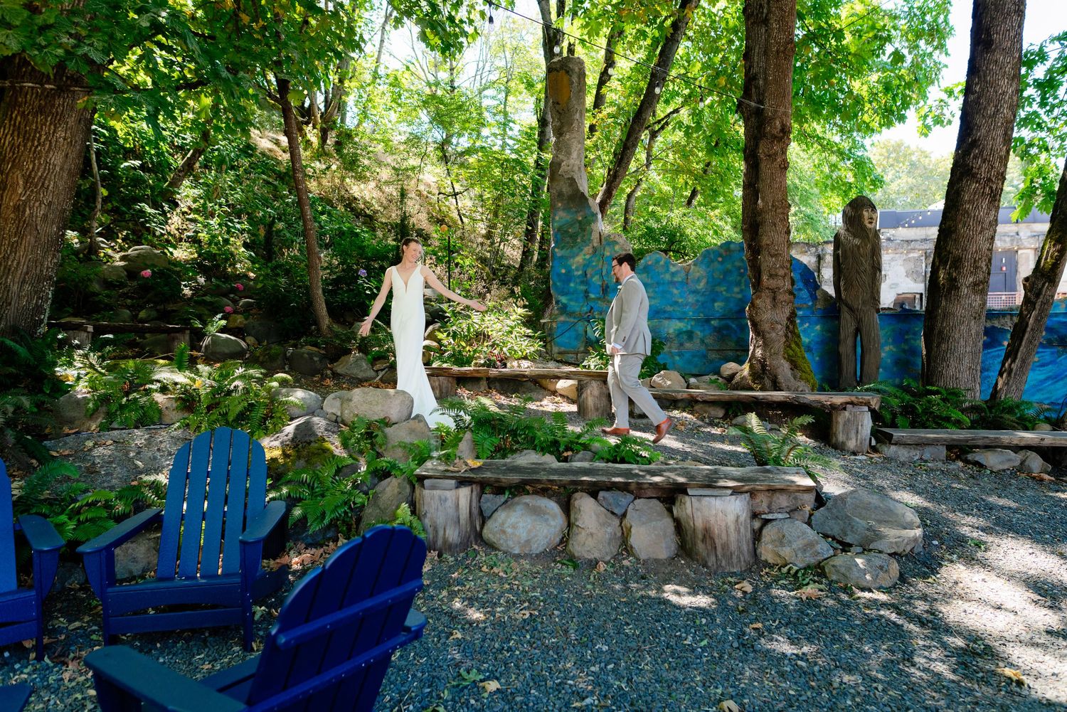 Couple walks through a lush garden with blue Adirondack chairs and stone seating at an outdoor wedding venue.