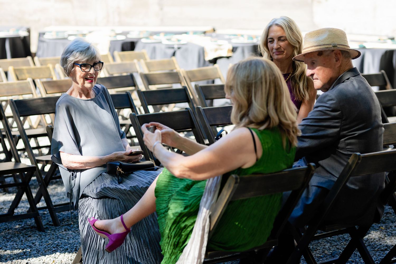 Four guests in elegant attire converse while seated on folding chairs at an outdoor wedding venue.