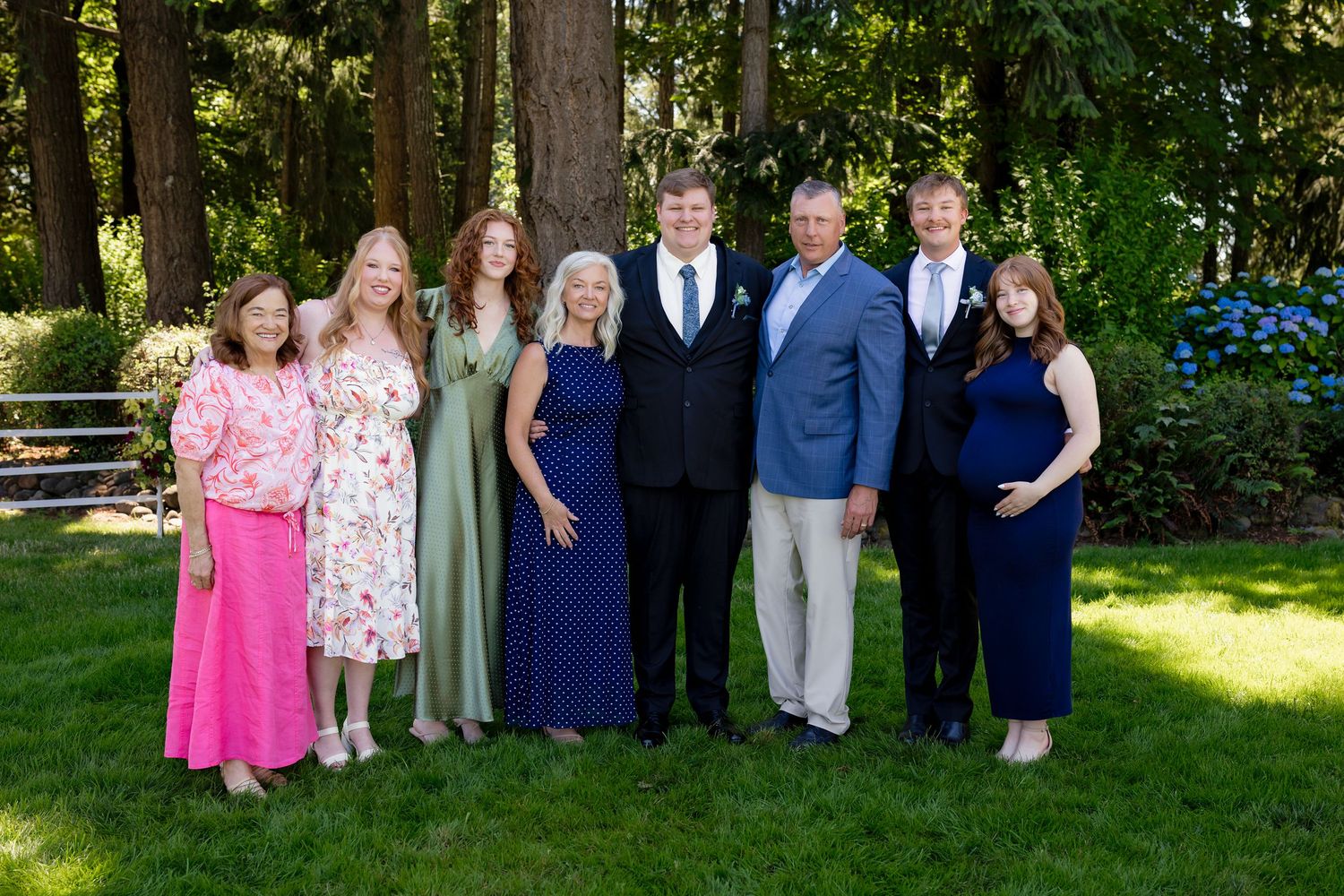 A family of seven poses together outdoors in formal attire among tall green trees on a sunny day.