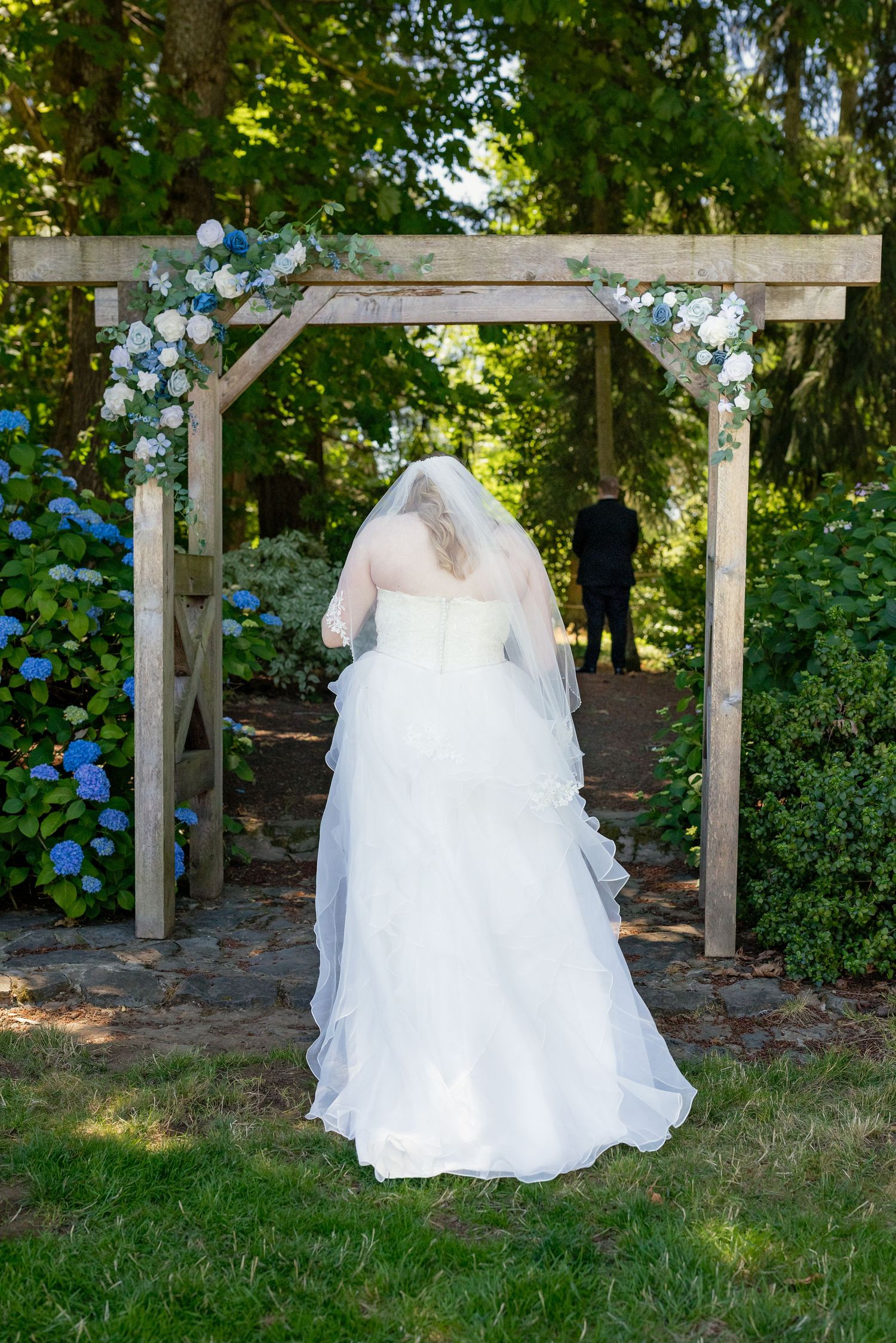 Bride in white gown and veil stands under floral wooden arch at outdoor garden wedding ceremony.