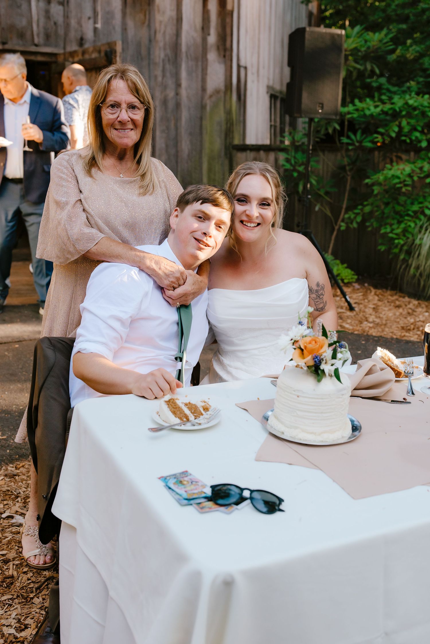 Bride in strapless white dress poses with teen boy and older woman near wedding cake.