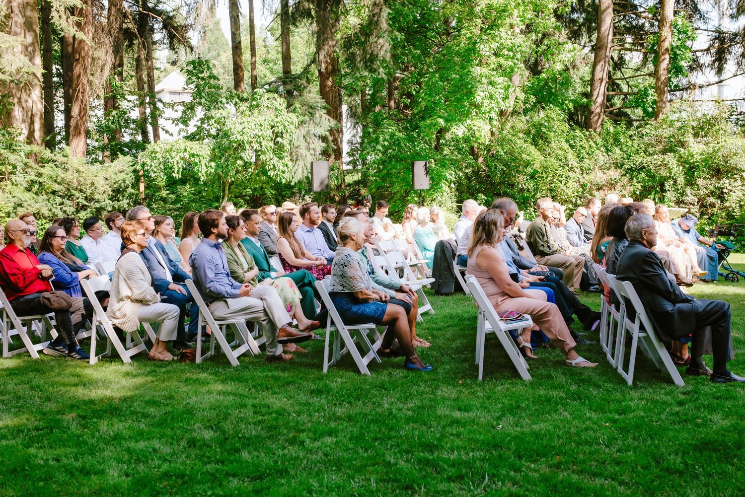 Guests seated on white chairs at an outdoor garden wedding ceremony surrounded by lush green trees.