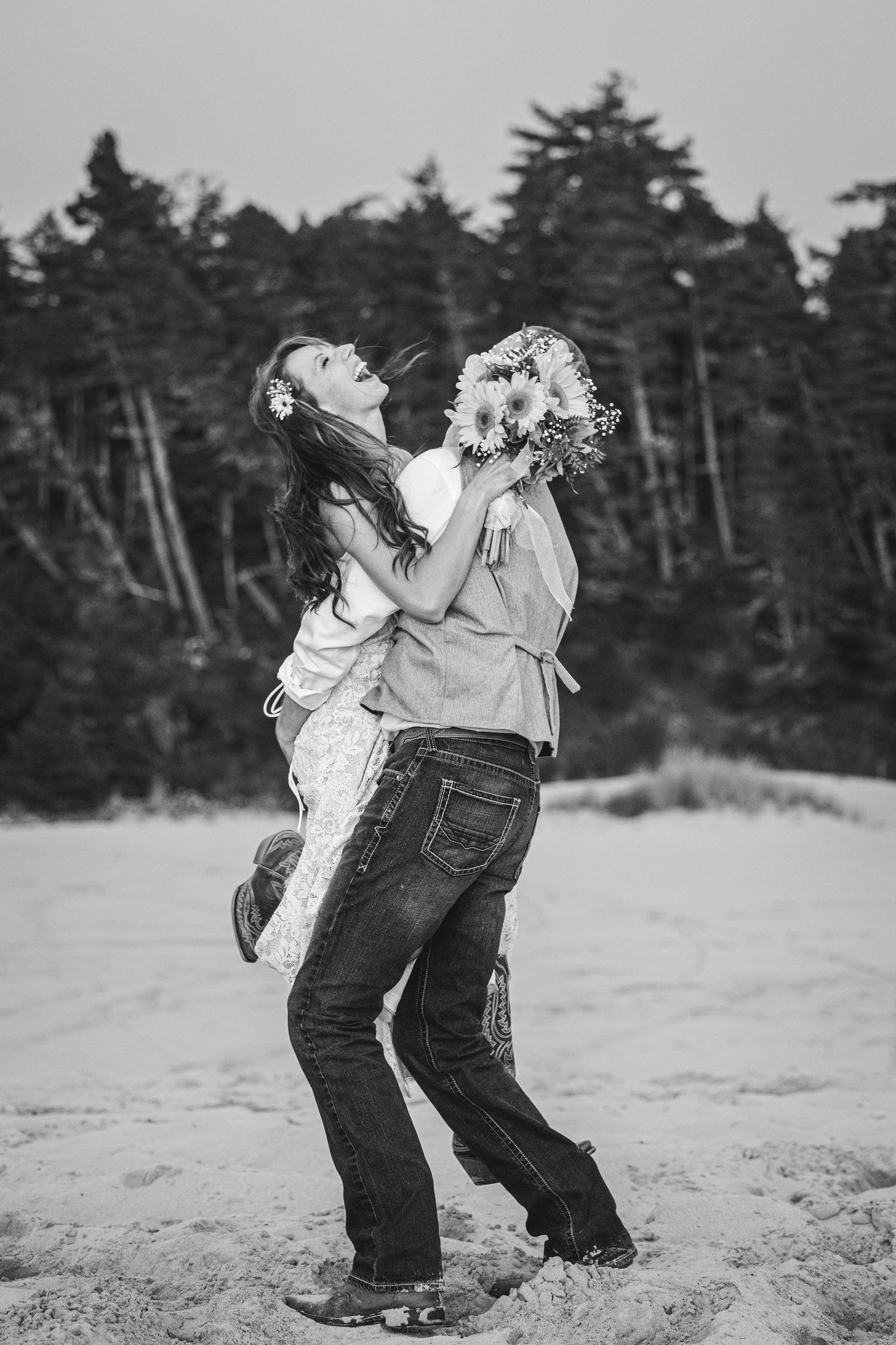 Black and white photo of a couple embracing on a sandy beach, woman holding sunflowers, trees in background.
