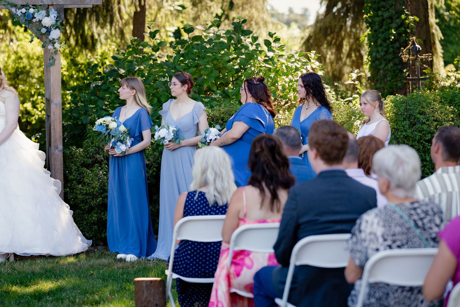 Bridesmaids in blue dresses standing at outdoor wedding ceremony with seated guests.