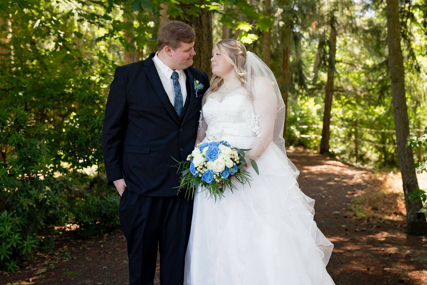 Bride and groom share a romantic moment in a lush forest setting, bride holding a blue and white bouquet.