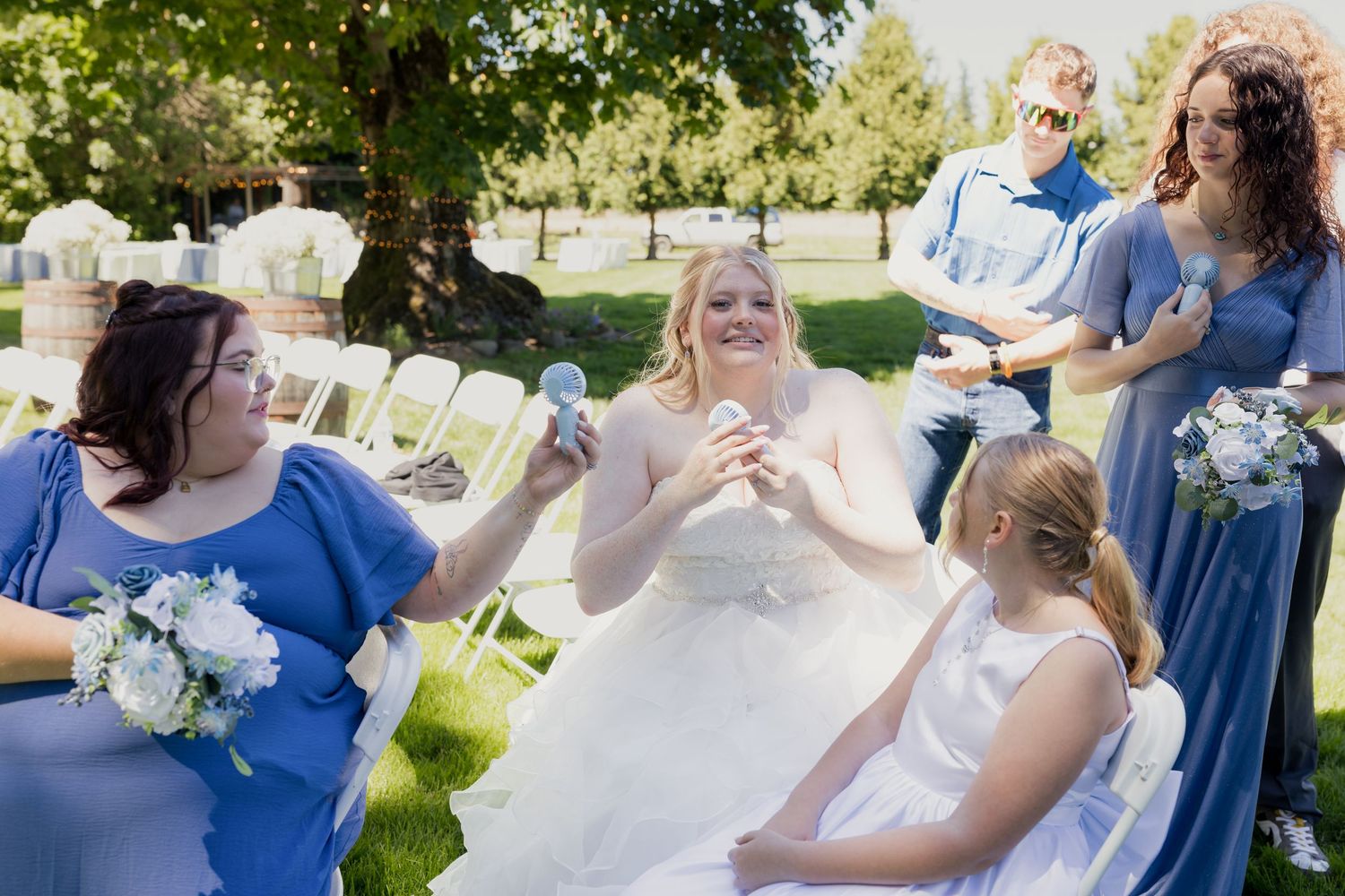 Bride in white gown laughs with bridesmaids in blue dresses during outdoor wedding ceremony on sunny day.