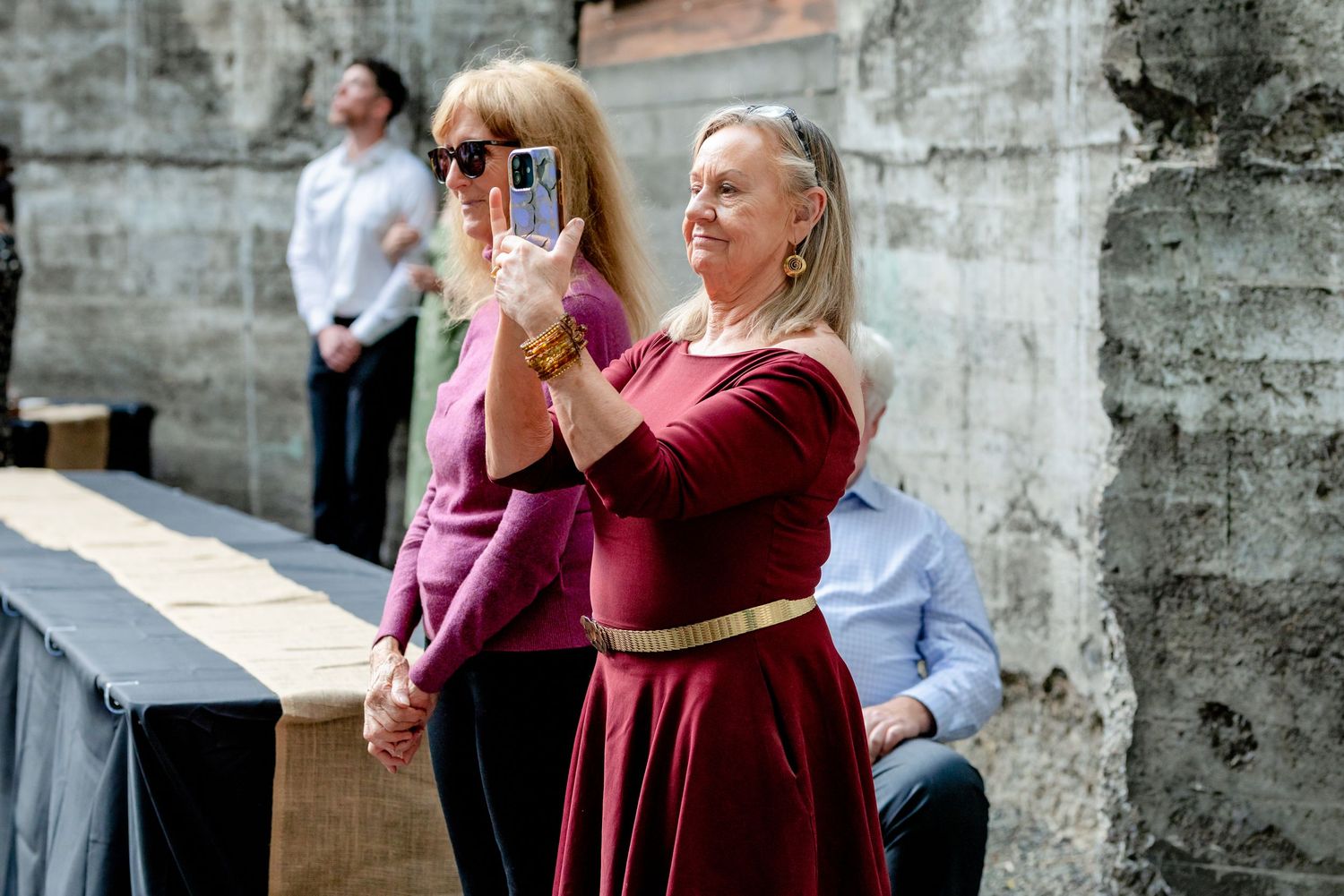 Two women in colorful dresses take photos on smartphones during an outdoor wedding ceremony.