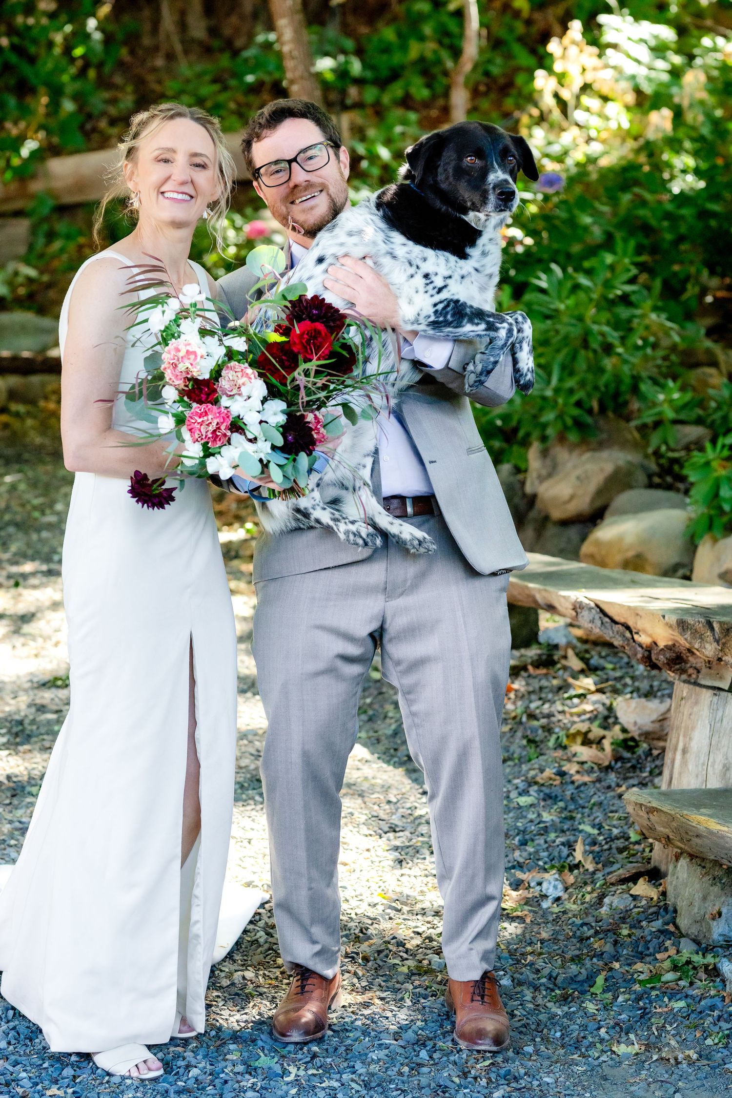 Happy bride holding floral bouquet and groom in gray suit pose with their black spotted dog outdoors.