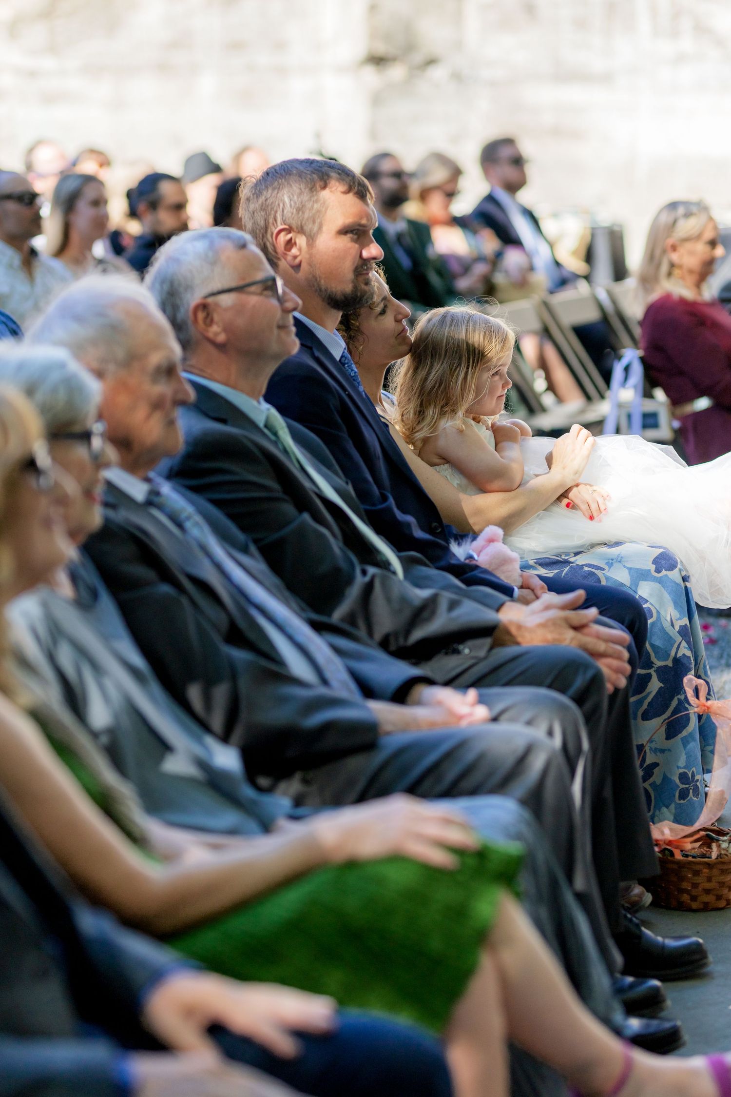 Guests seated in rows at an outdoor wedding ceremony, including a young girl in a white dress and men in suits.