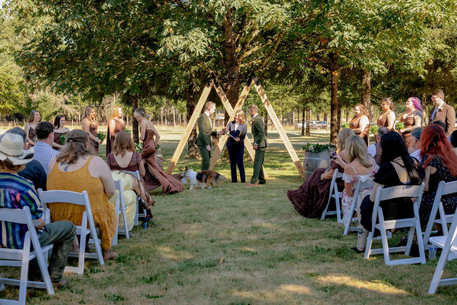 Outdoor wedding ceremony with guests seated in white chairs facing couple at triangular wooden arch.