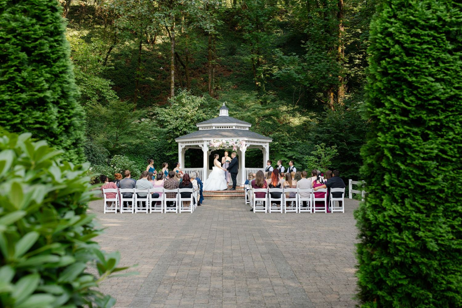 Outdoor wedding ceremony at a white gazebo surrounded by lush greenery with seated guests.