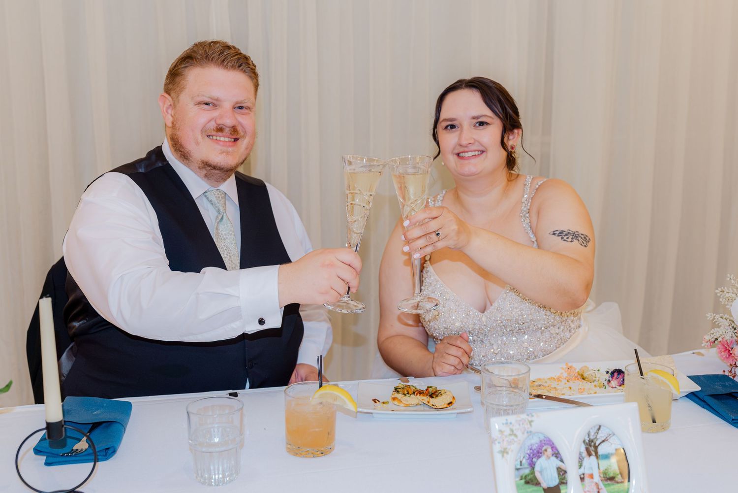 Bride and groom toast with champagne flutes while seated at sweetheart table at reception.