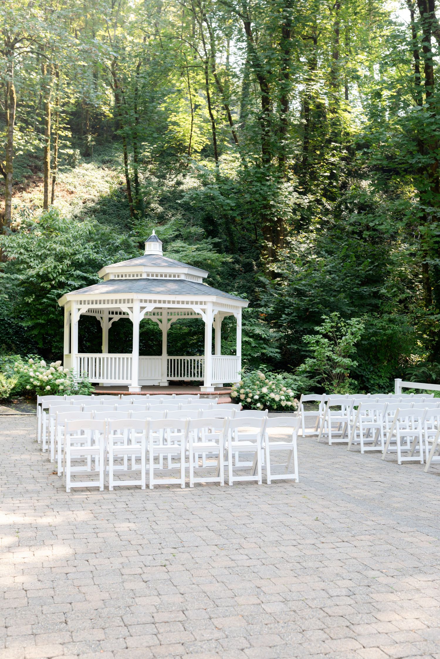 White gazebo wedding ceremony setup with folding chairs on brick patio surrounded by lush green forest.