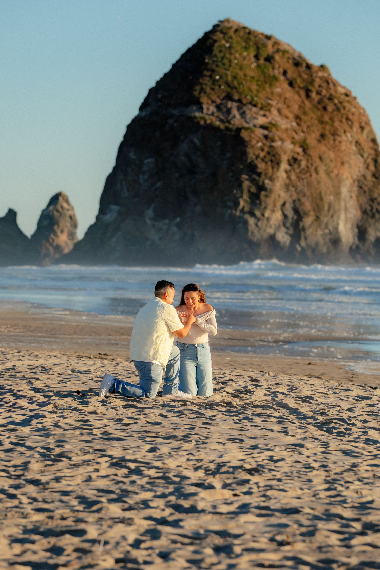 Man proposes on one knee to woman on sandy beach with iconic Haystack Rock at Cannon Beach, Oregon at sunset.