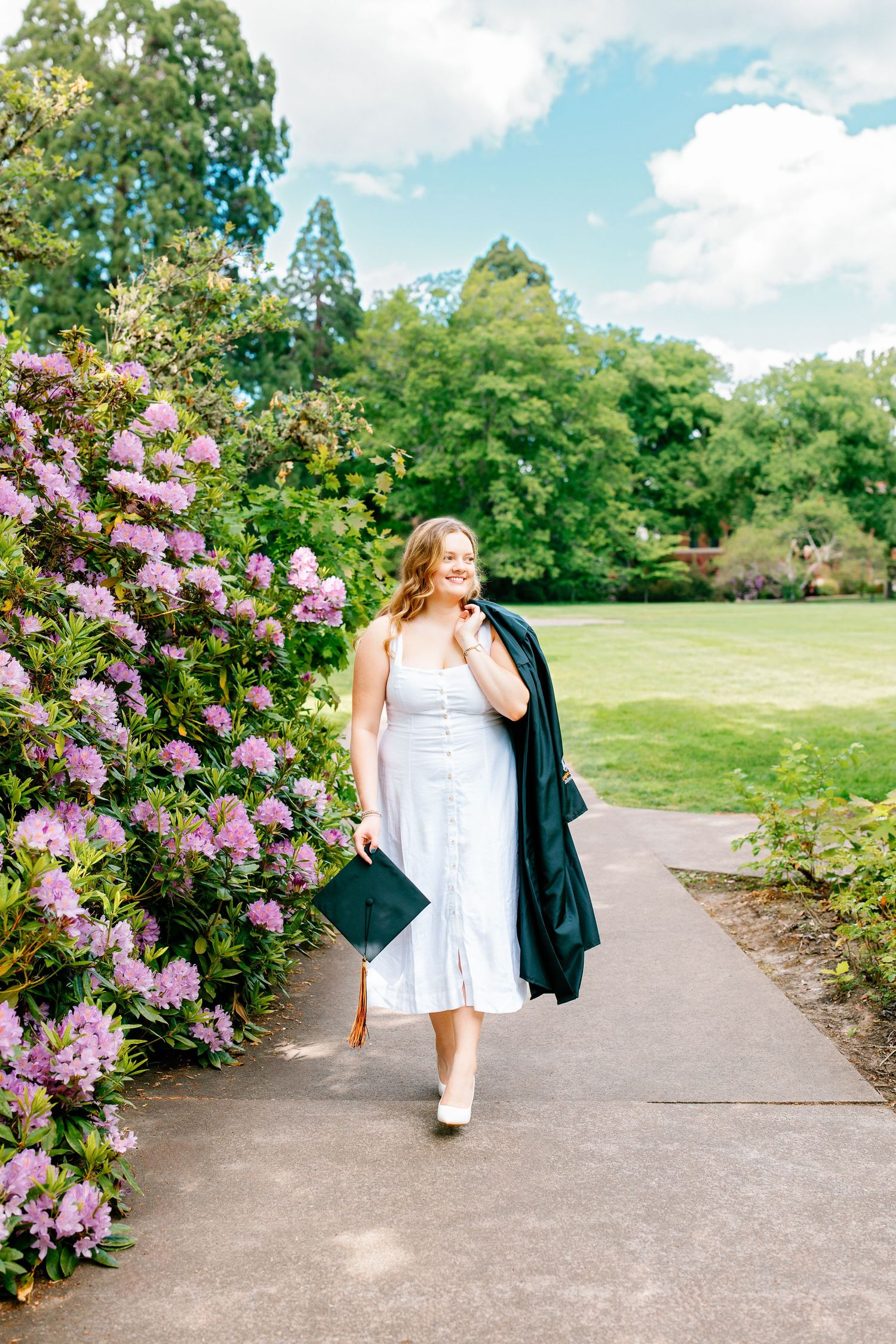 Young woman in white dress holding graduation cap and gown walks along garden path with blooming purple flowers.
