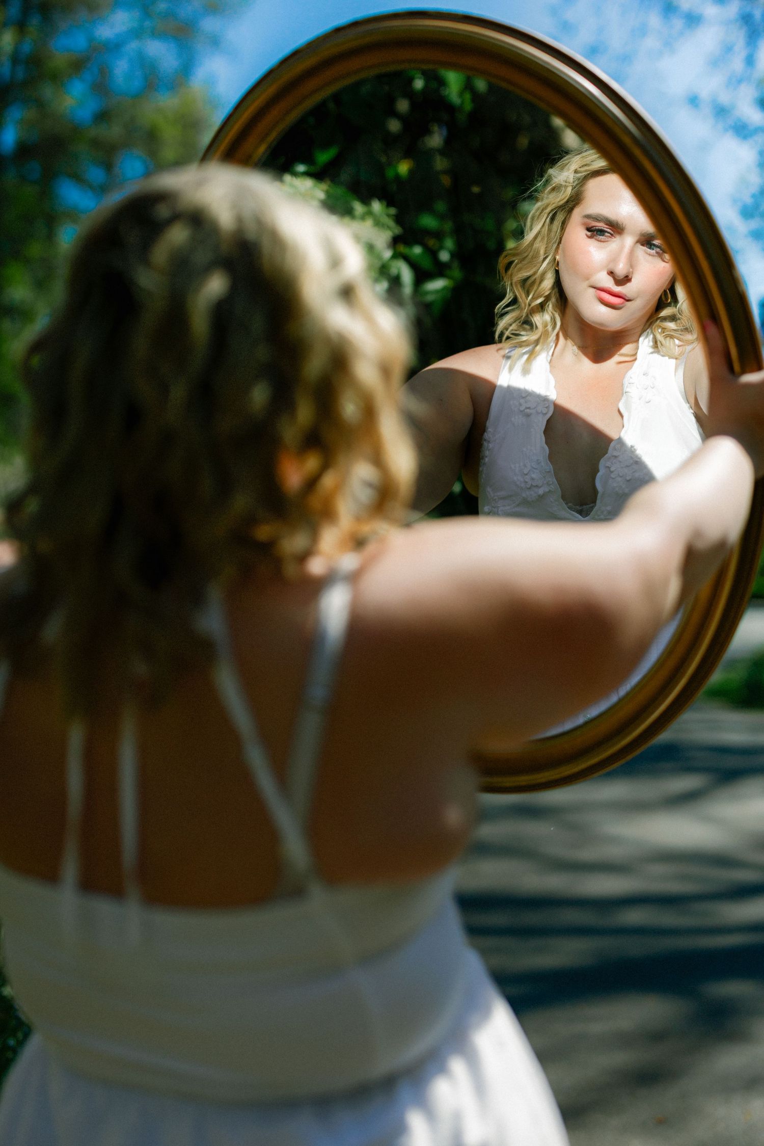 Blonde woman in white dress holding a round mirror outdoors, her reflection gazing back in natural sunlight.