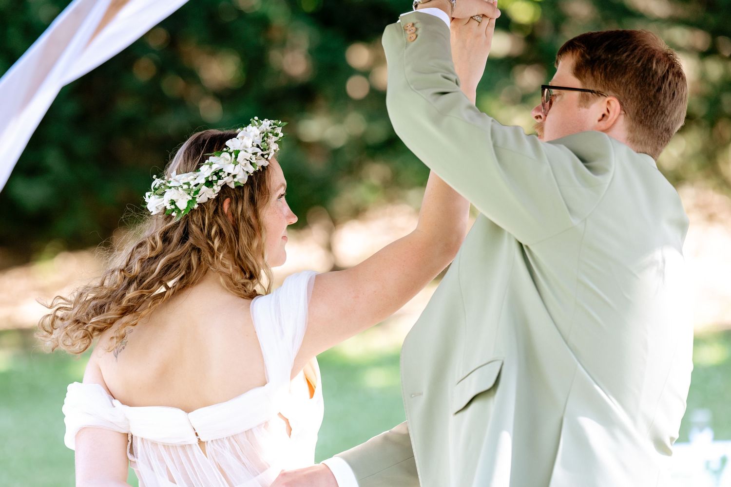 Bride in floral crown and groom in sage suit dancing outdoors at a romantic garden wedding ceremony.