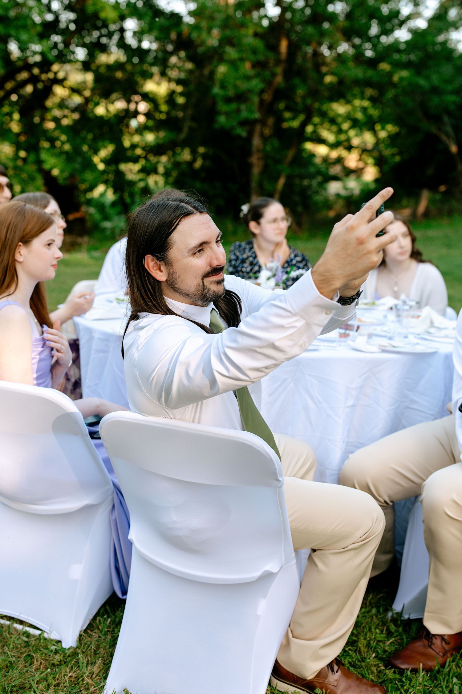 Man with long hair gestures at outdoor garden wedding reception seated at white draped table with guests.