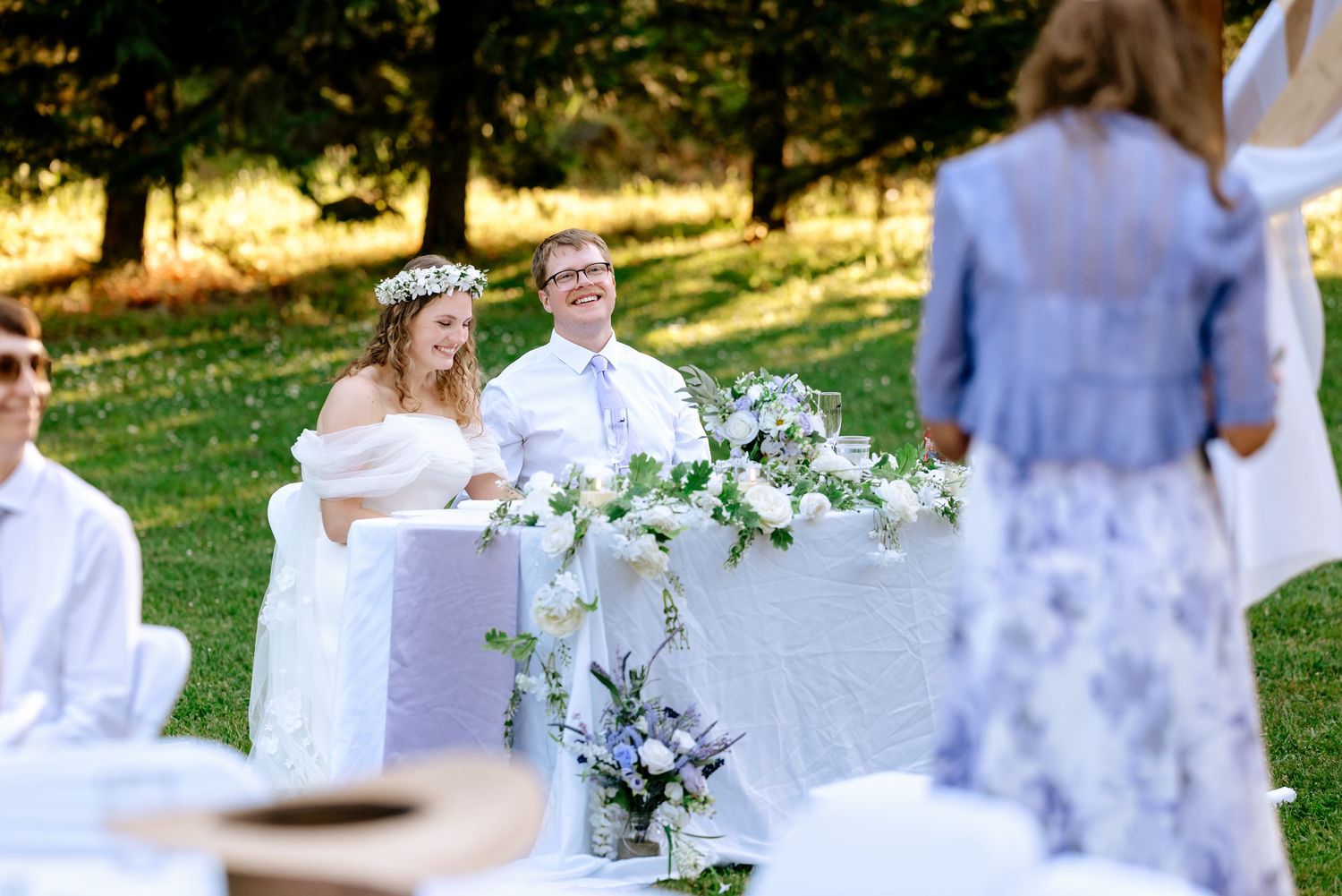 Bride and groom smile at sweetheart table decorated with white flowers during outdoor garden reception.