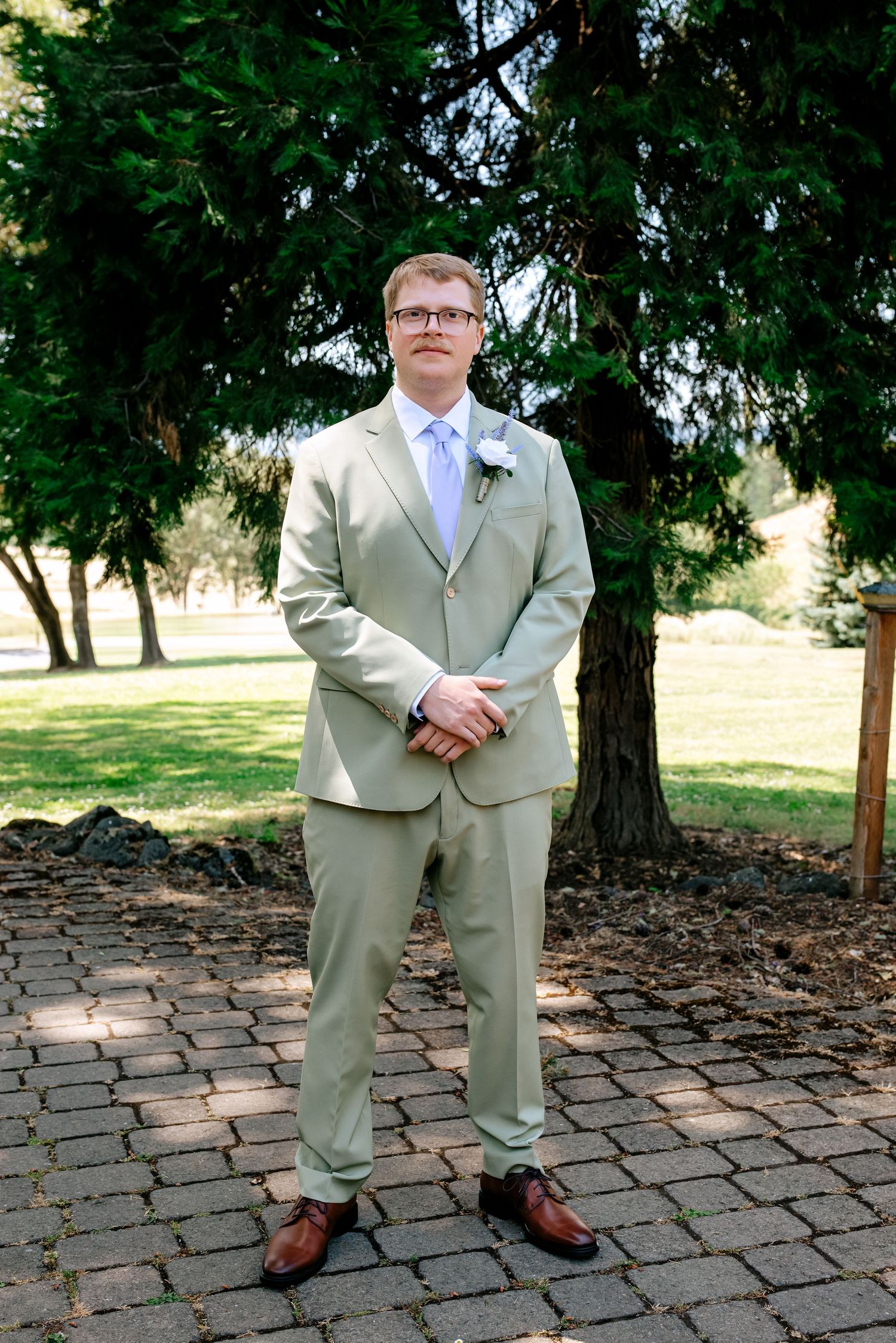 Groom in light khaki suit with lavender tie and boutonniere standing on brick path before wedding ceremony.