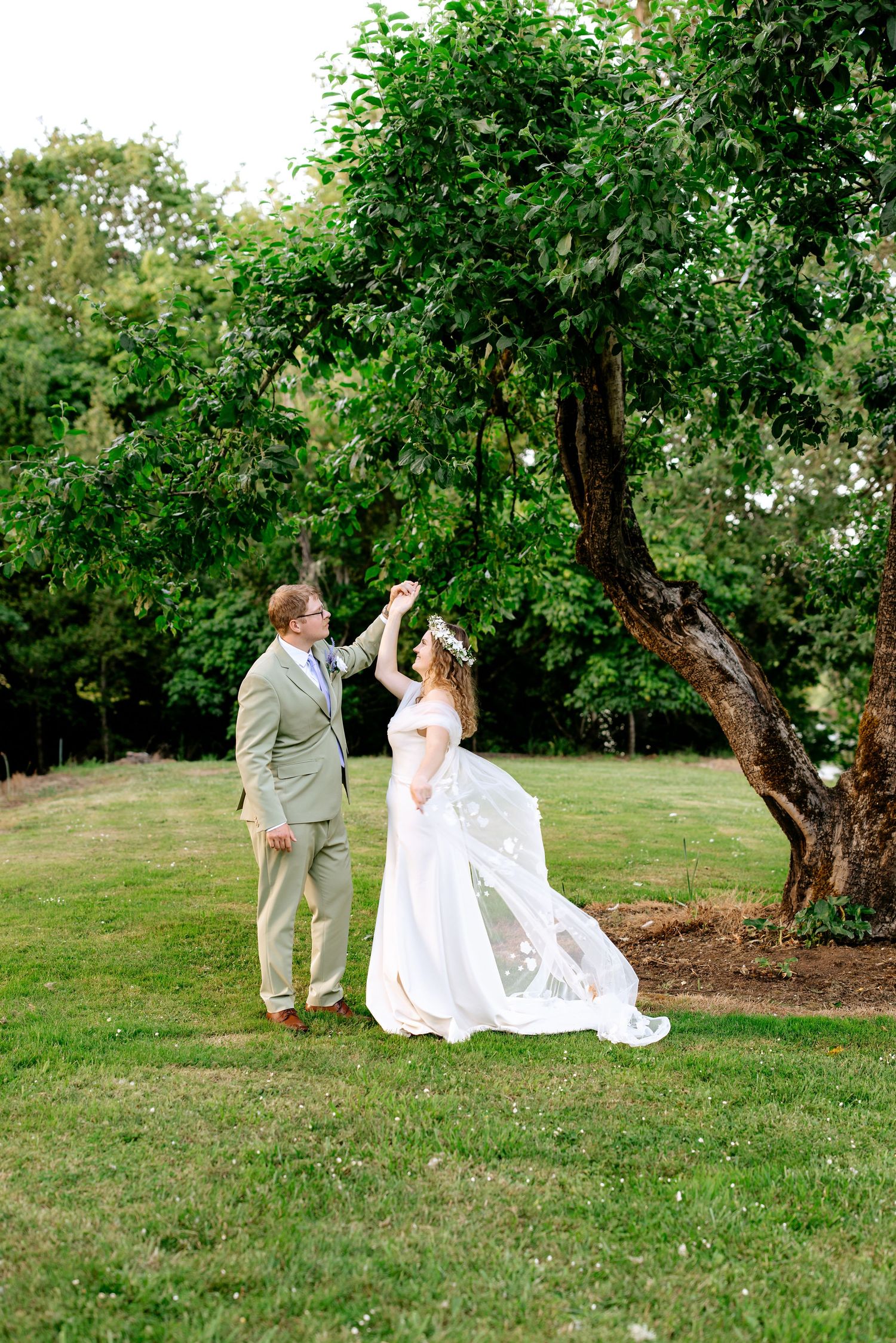 A groom in a tan suit twirls his bride in a white gown and floral crown outdoors on a green lawn near a large tree.