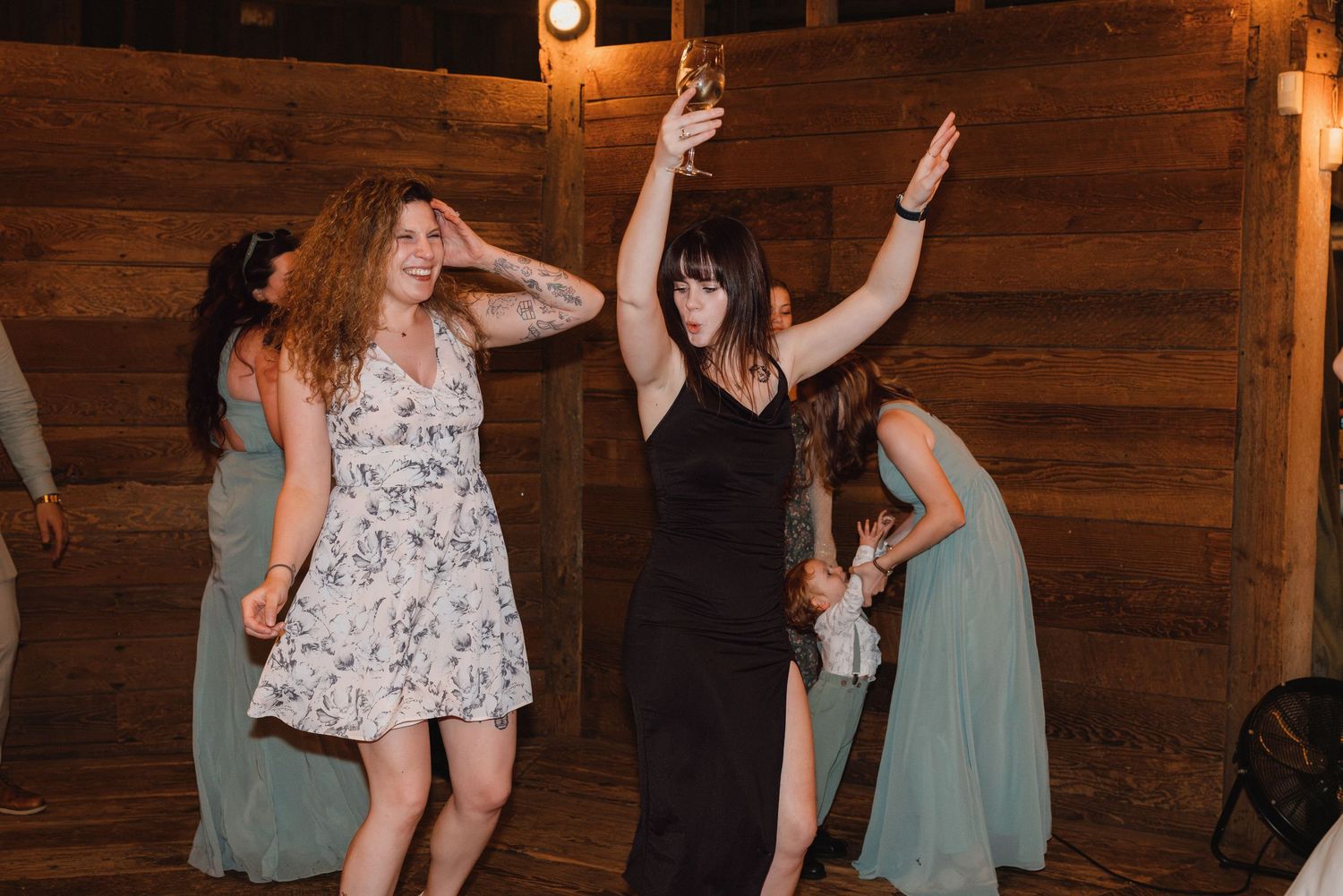 Two women in dresses dance energetically at rustic barn wedding reception under warm lighting.