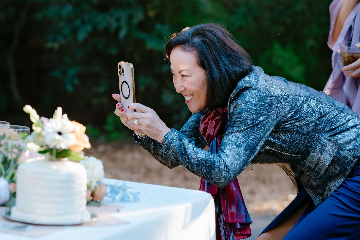 Woman smiling while photographing wedding cake with smartphone at outdoor reception table.