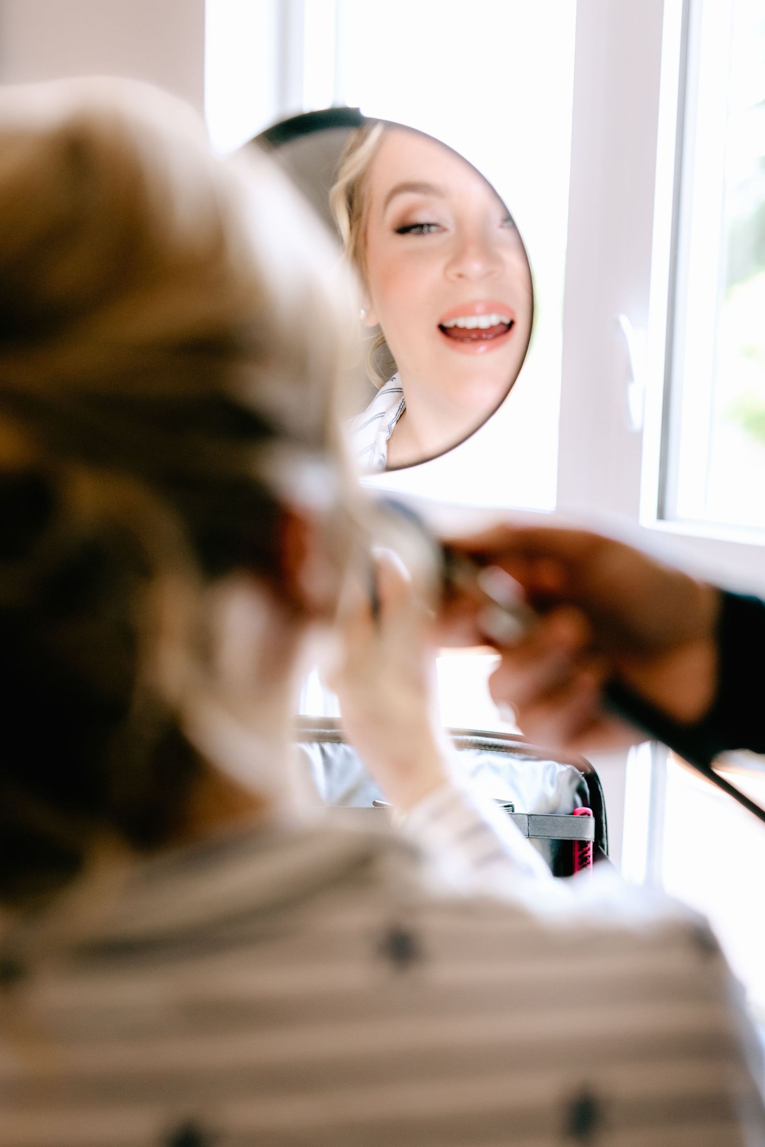 Bride smiling at her reflection in a round mirror while getting makeup applied on her wedding day.