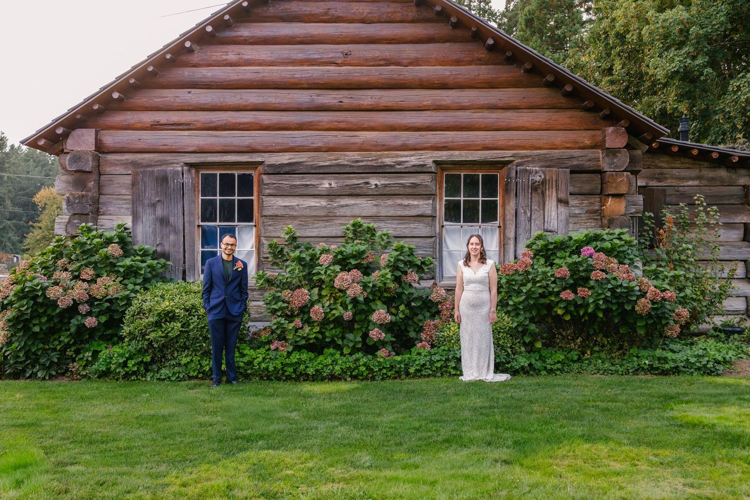 A rustic wooden barn with pink roses growing along its weathered exterior sits in a garden setting.