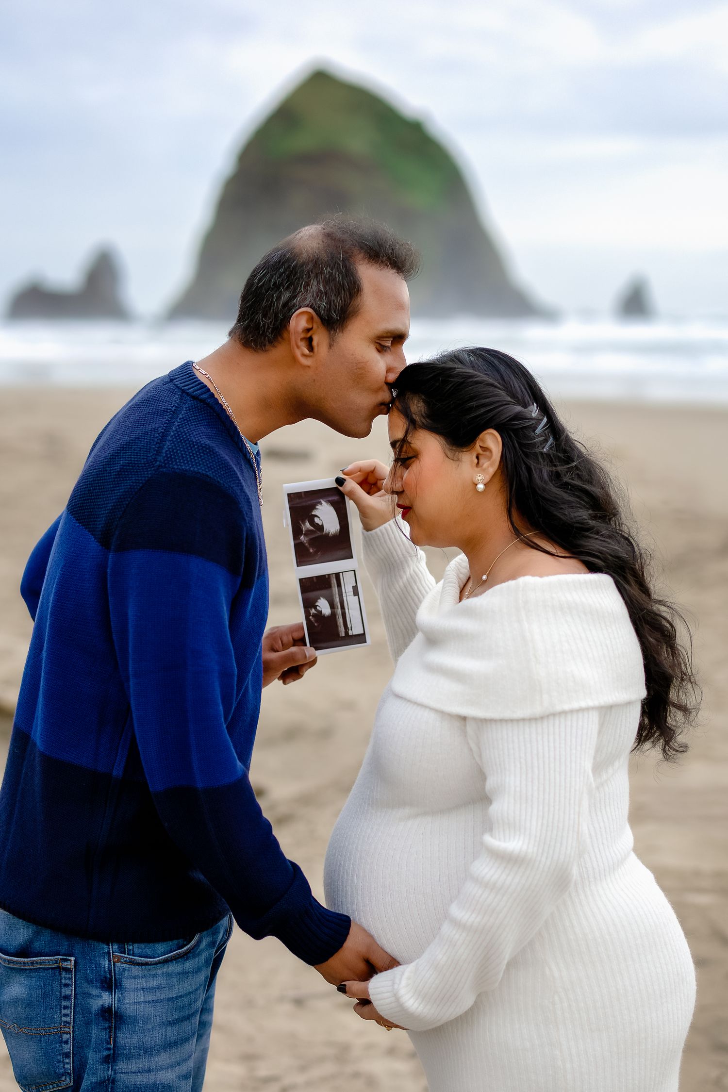 A couple looks at ultrasound photos together on a misty beach with rock formations in the distance.