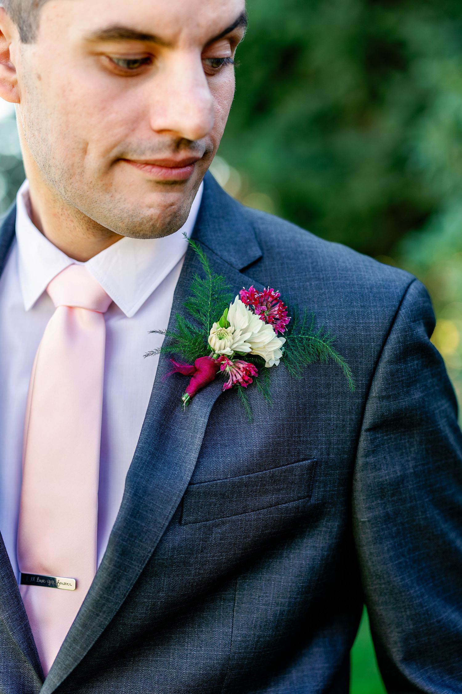 Groom wearing charcoal suit with pink tie and burgundy boutonniere in outdoor garden setting.