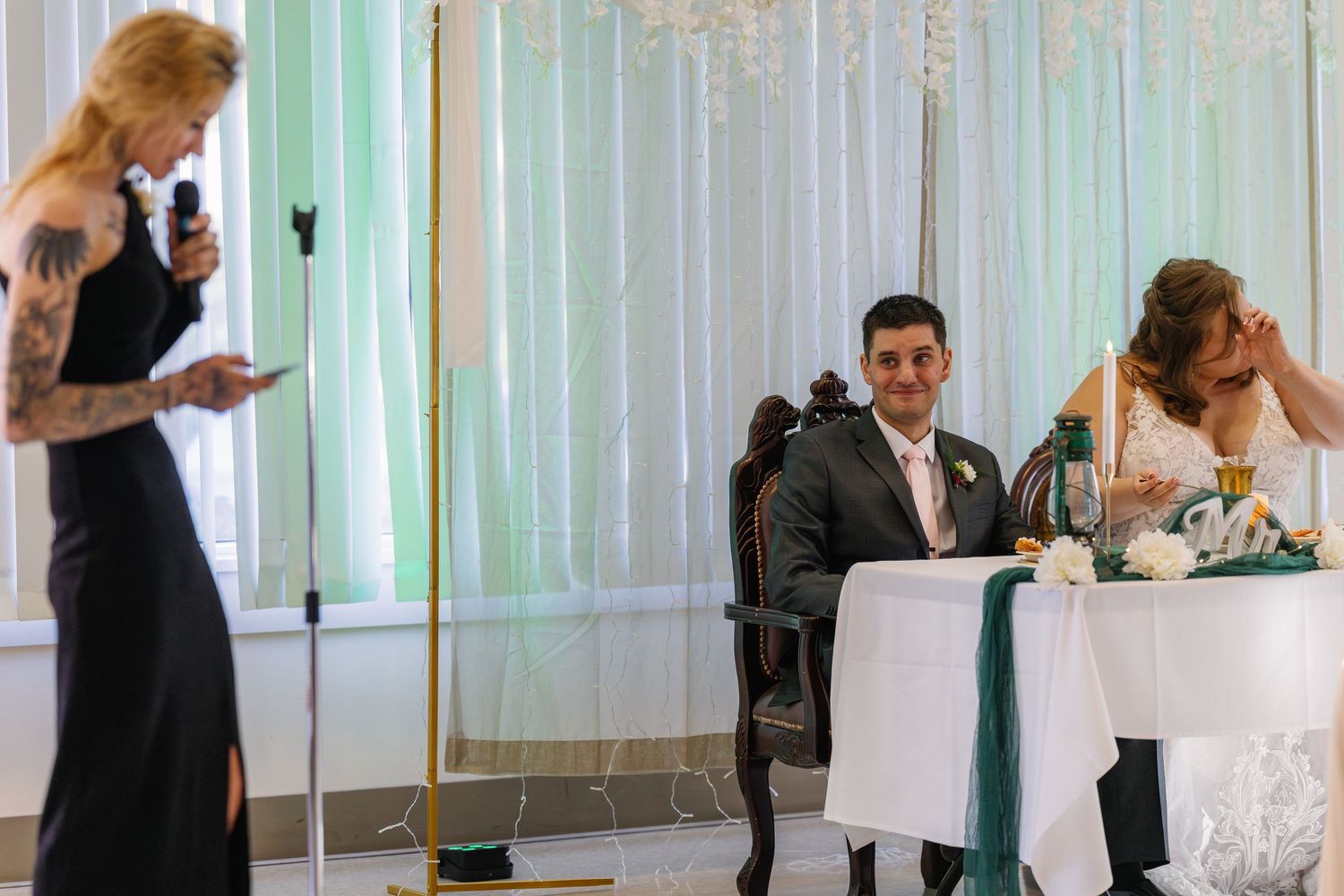 Wedding reception scene with white backdrop and head table during speeches.