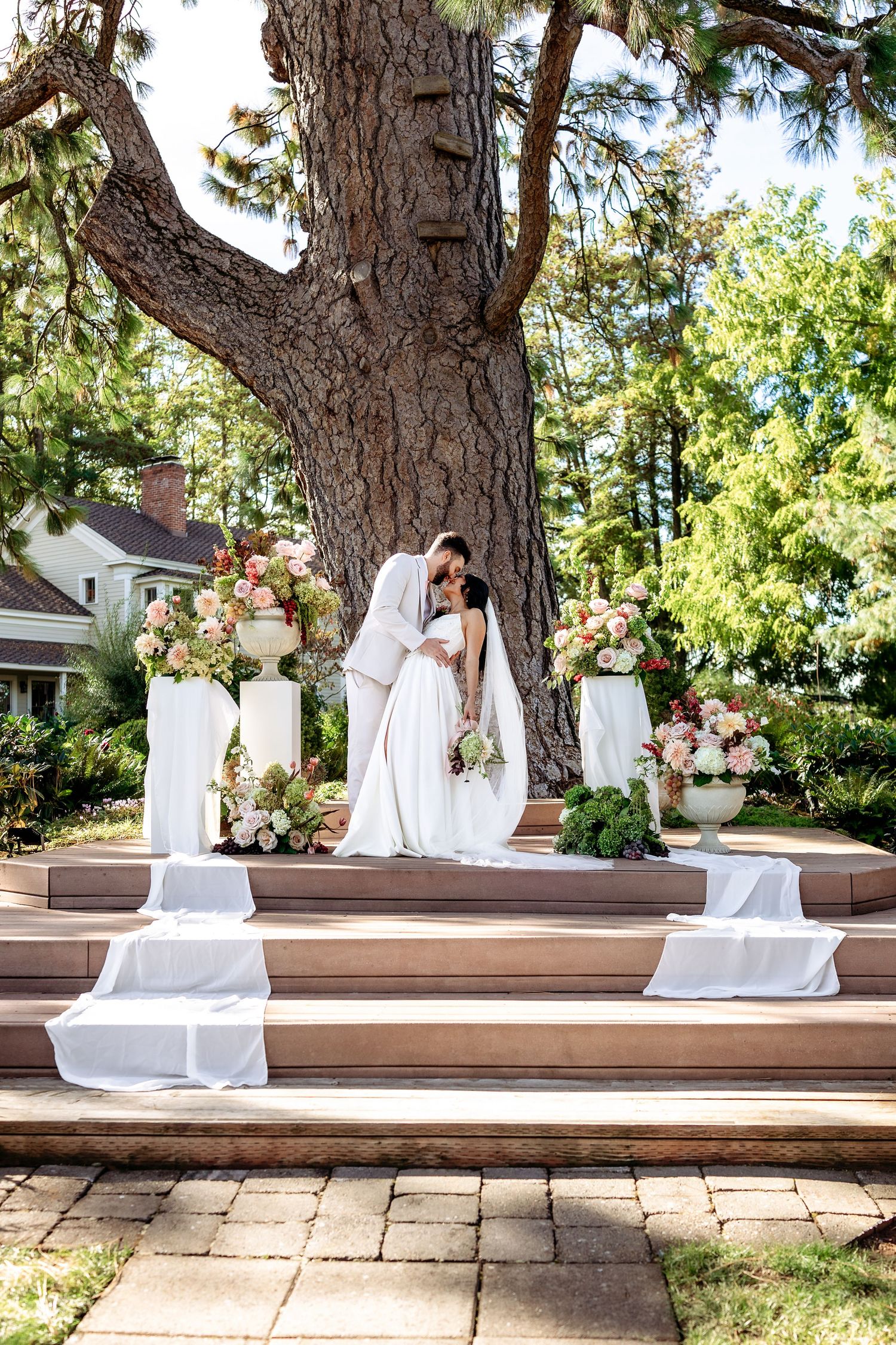 A couple shares an intimate dance on a wooden deck beneath a large tree at their outdoor wedding.