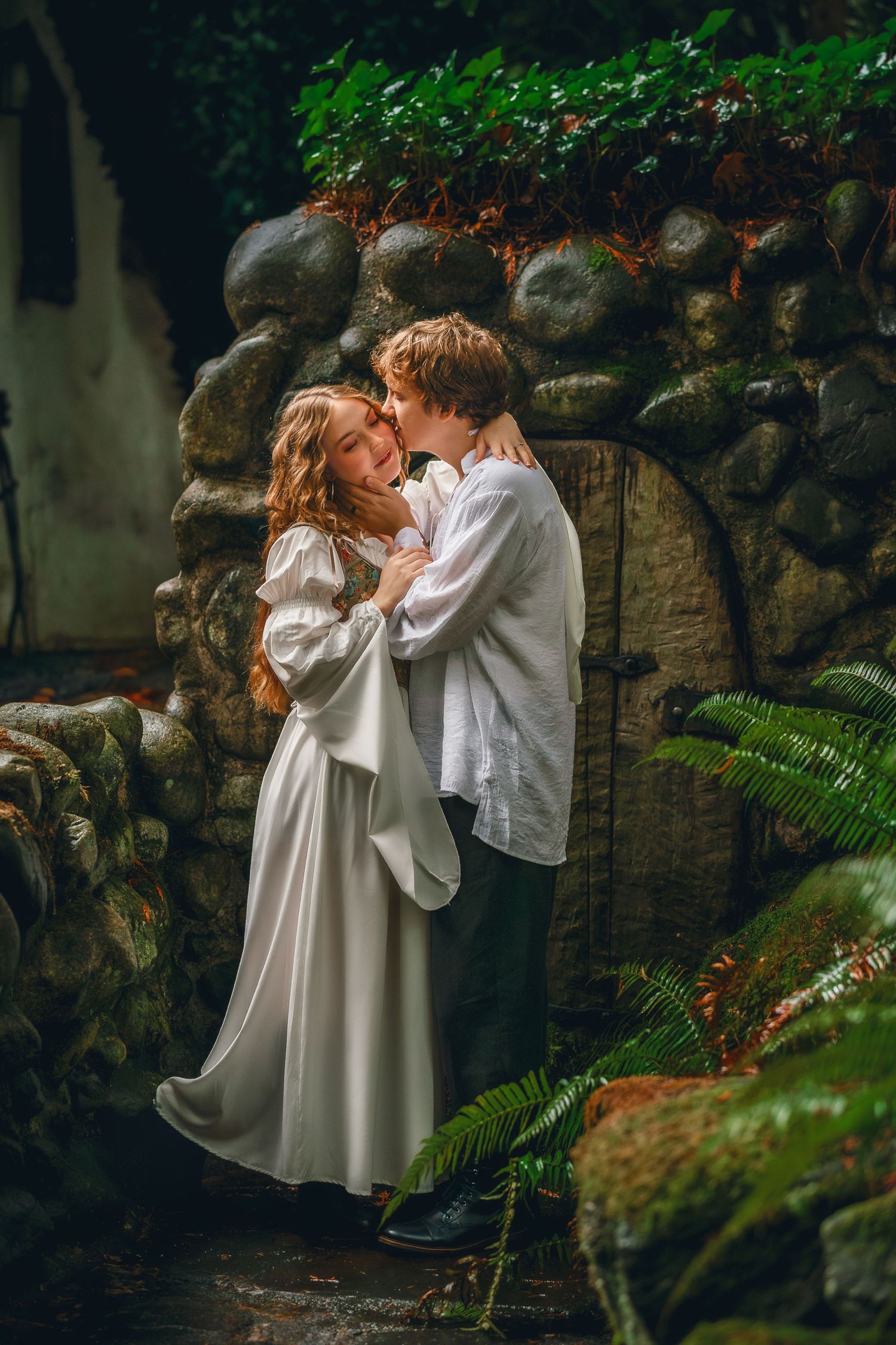 A romantic couple in white clothing embraces against a stone wall surrounded by lush greenery.