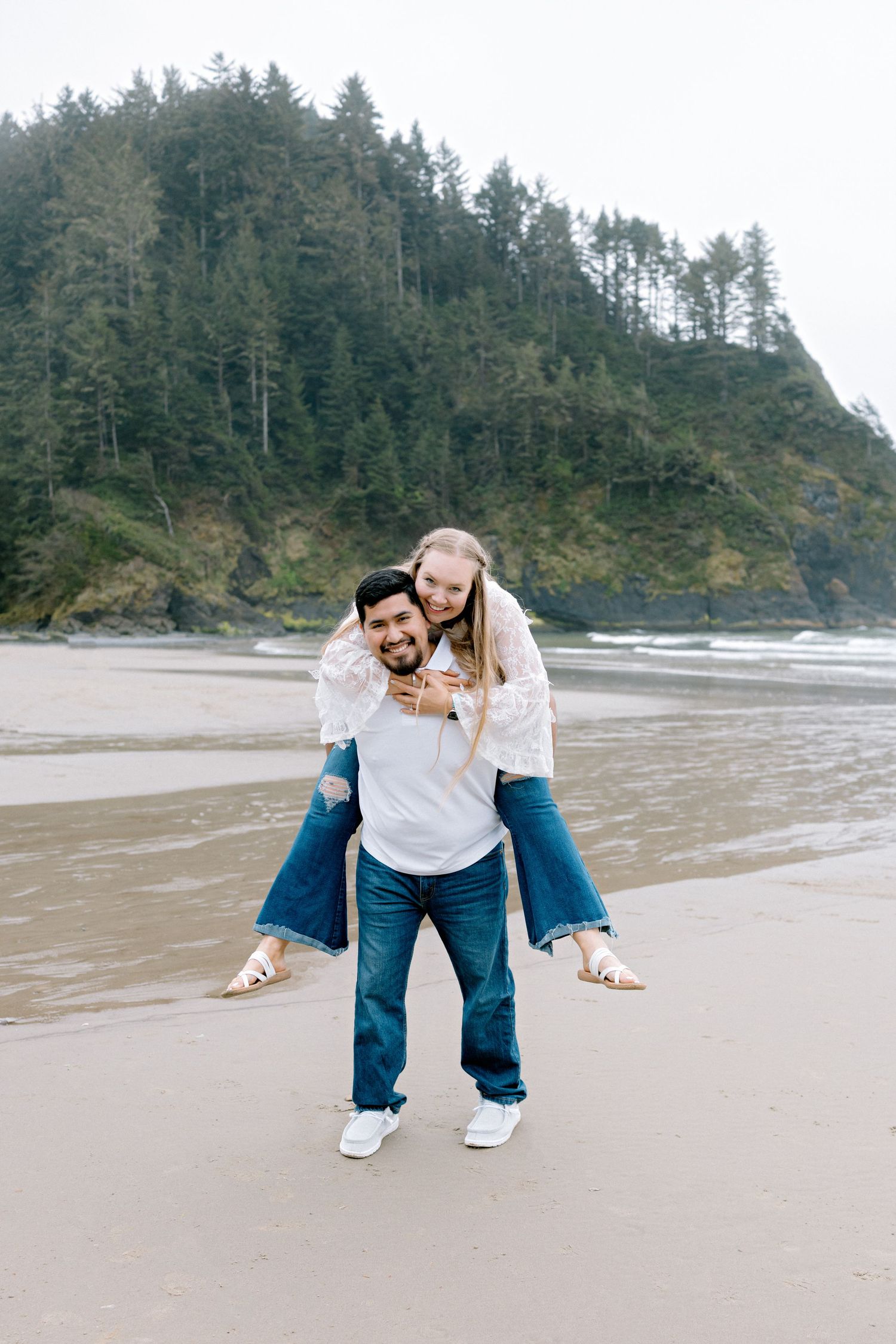 Man giving woman a piggyback ride on a sandy beach with forested cliffs in the background.