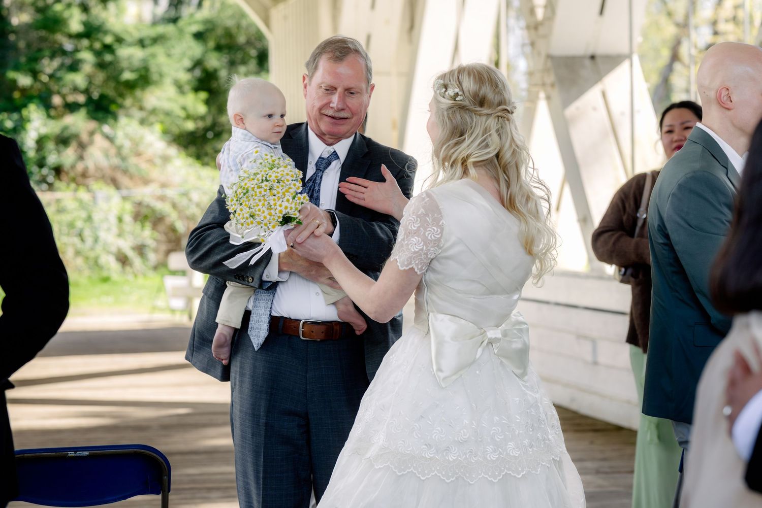 Father in gray suit holds baby with bouquet while greeting bride in white wedding gown.