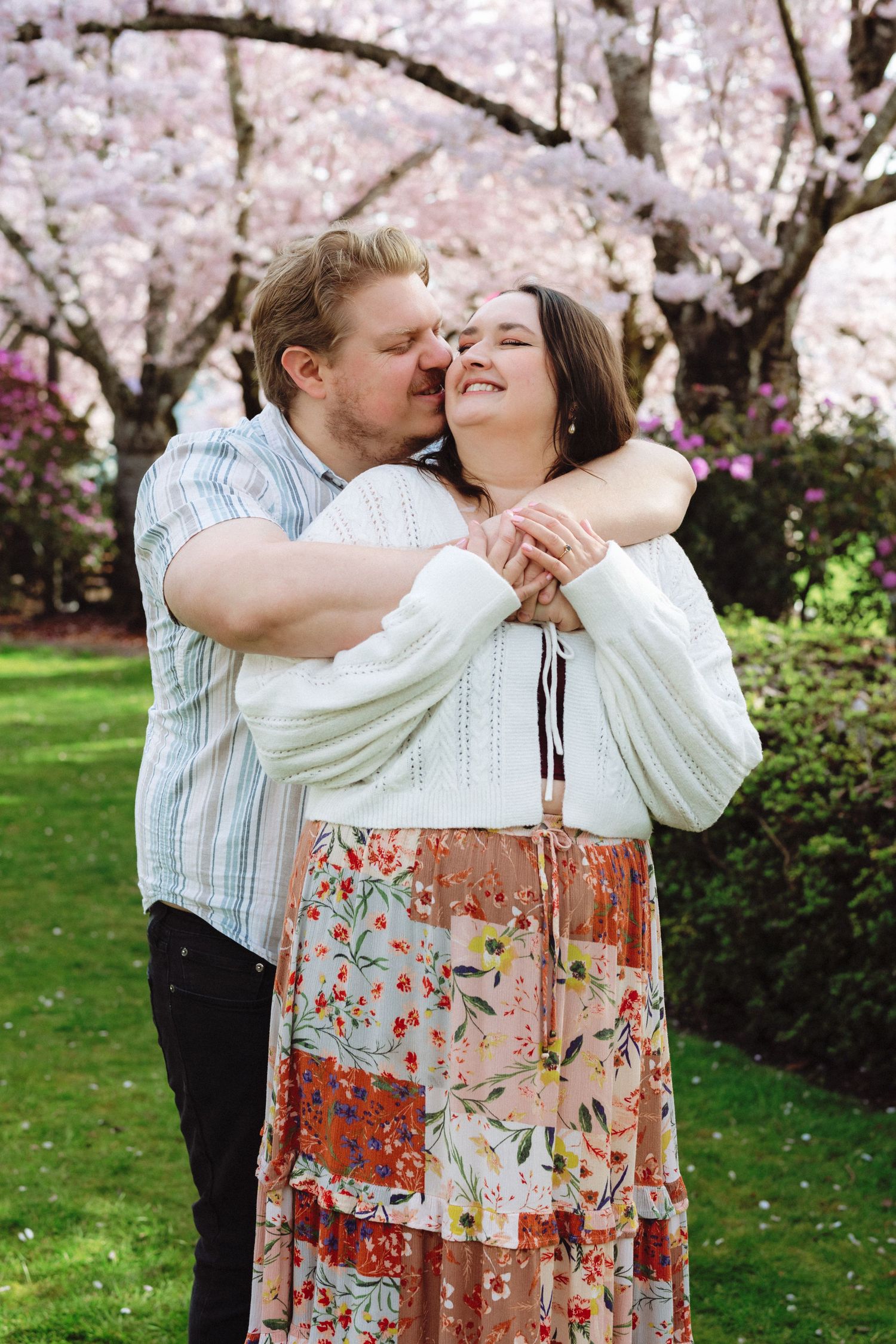 A romantic couple embraces under blooming cherry blossom trees in a spring garden setting.
