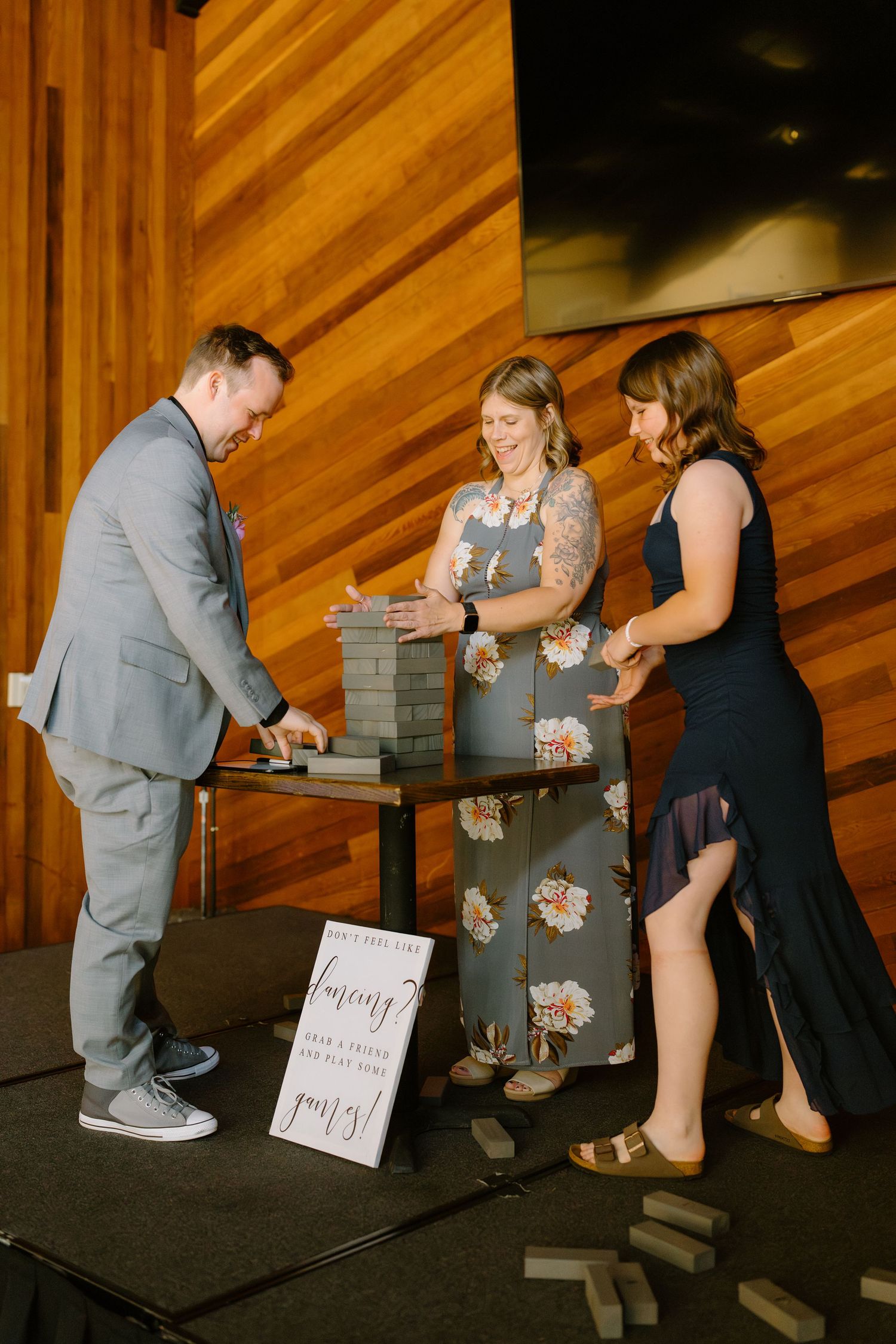 Three guests play a giant Jenga game at an elegant indoor wedding reception with warm wood panel walls.